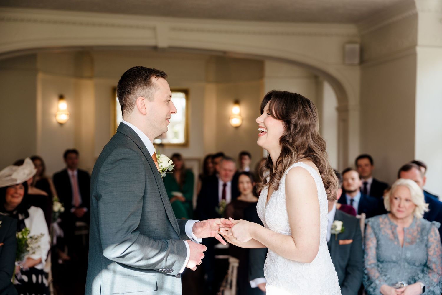 Laughing bride and groom exchange rings at the altar during their wedding ceremony with guests seated behind.