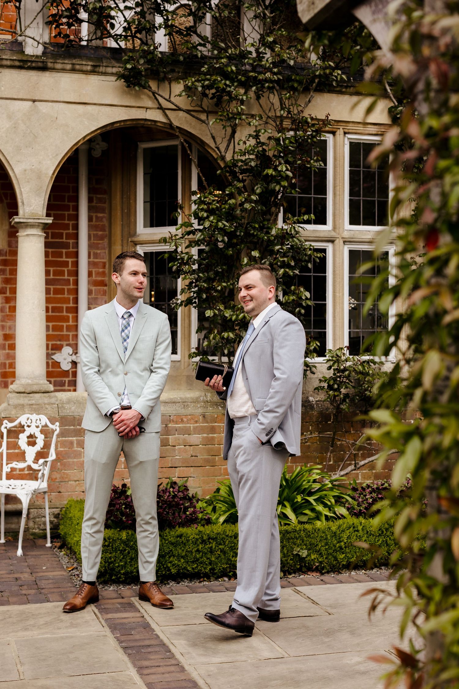 Two groomsmen in light grey suits conversing outside elegant manor house with arched stone facade.