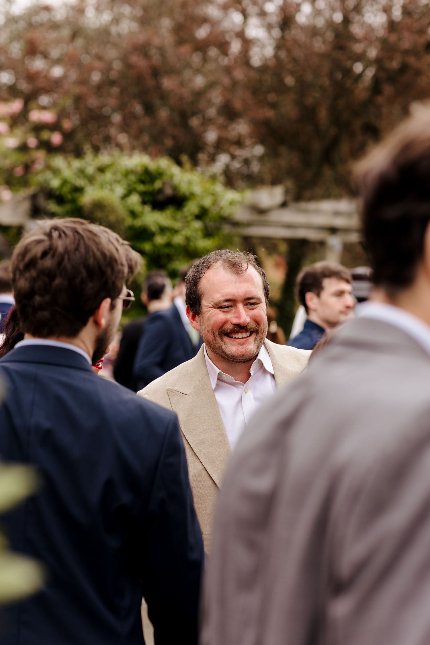 Smiling male wedding guest in beige blazer socializing outdoors at wedding reception.
