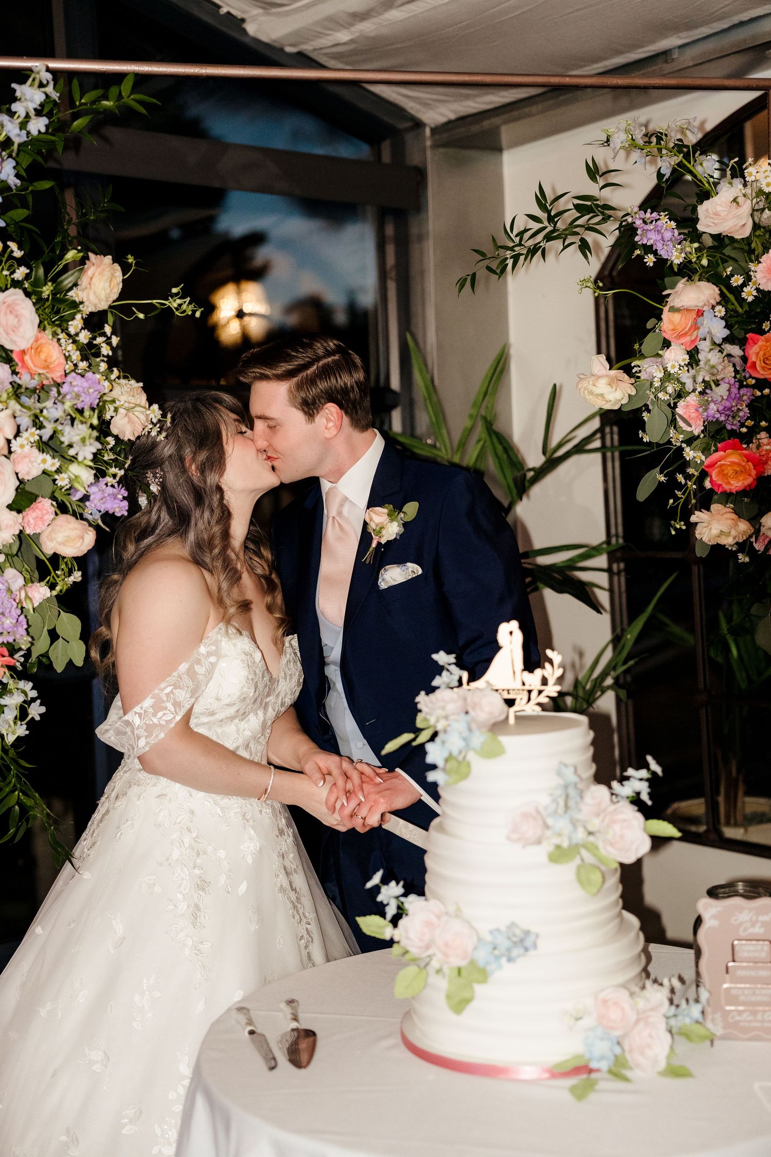Bride and groom kissing while cutting elegant white tiered wedding cake adorned with pastel flowers.