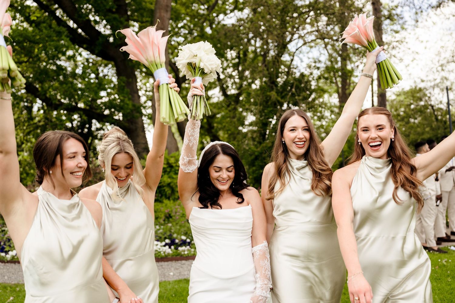 Five bridesmaids in ivory halter satin dresses celebrate outdoors raising bouquets with bride in white.