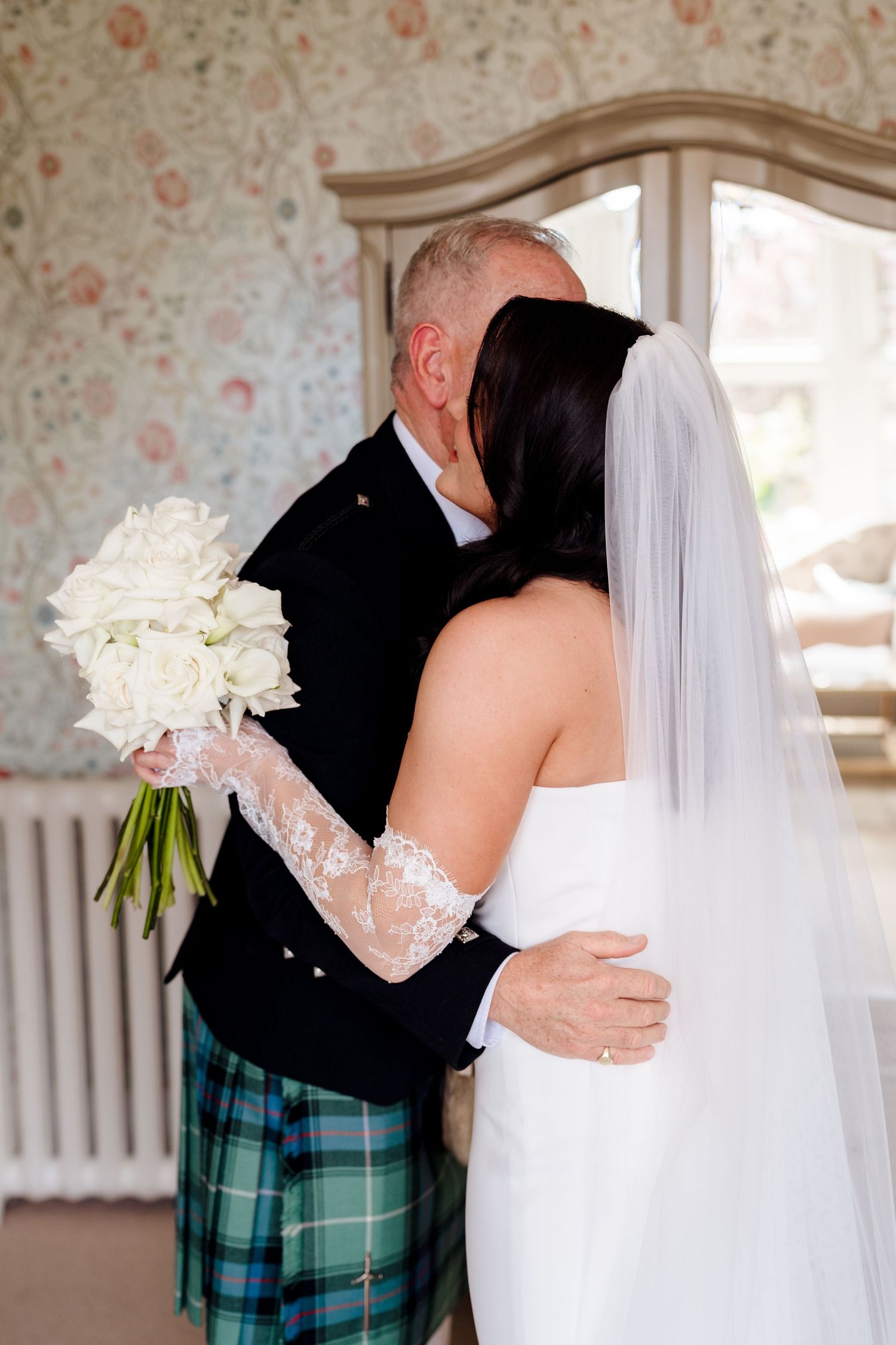 Bride and groom sharing a kiss, bride holds white bouquet, groom wears green tartan kilt, indoors with floral wallpaper.