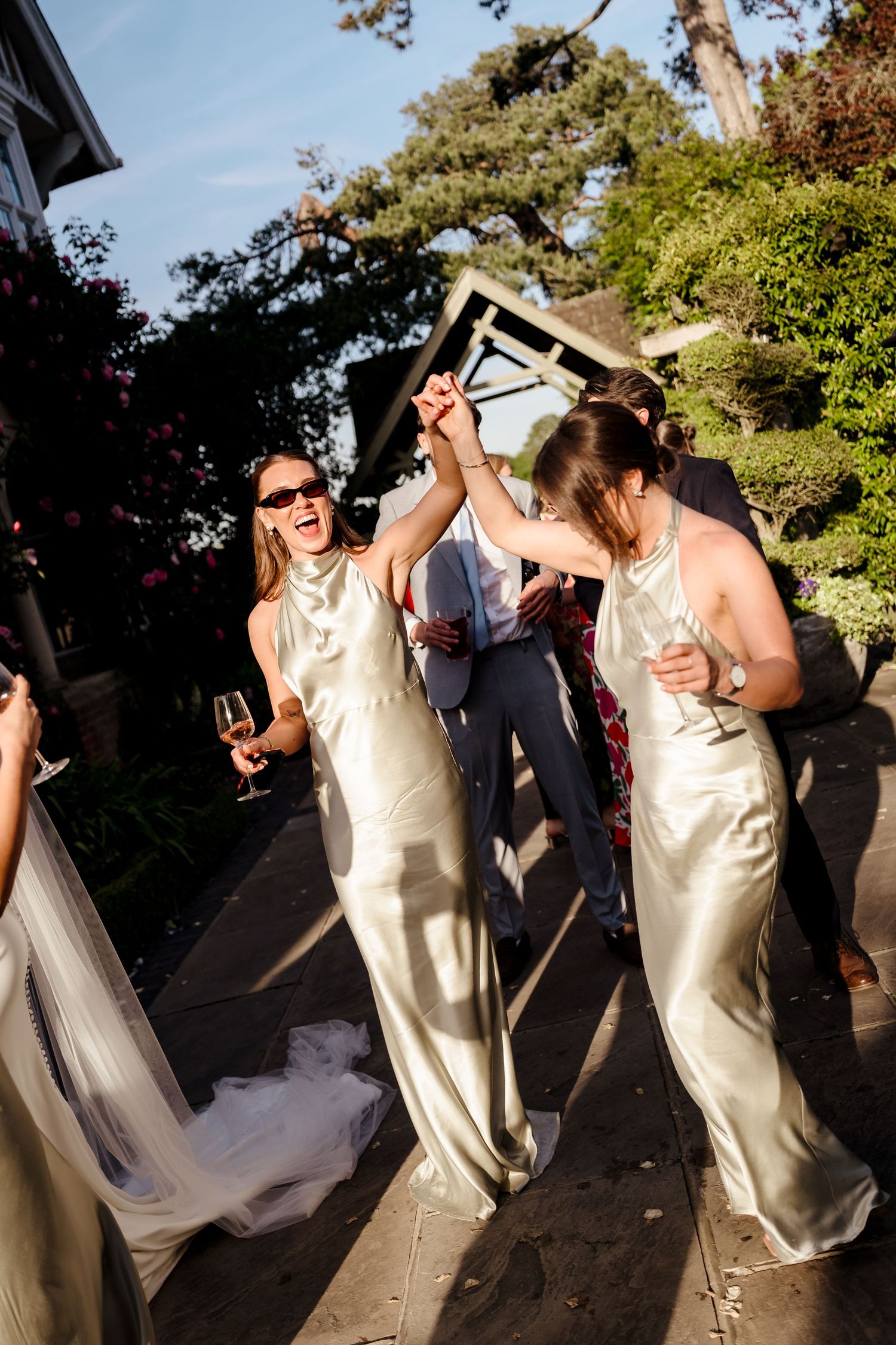 Bridesmaids in sage satin dresses dancing and celebrating outdoors in golden sunlight.