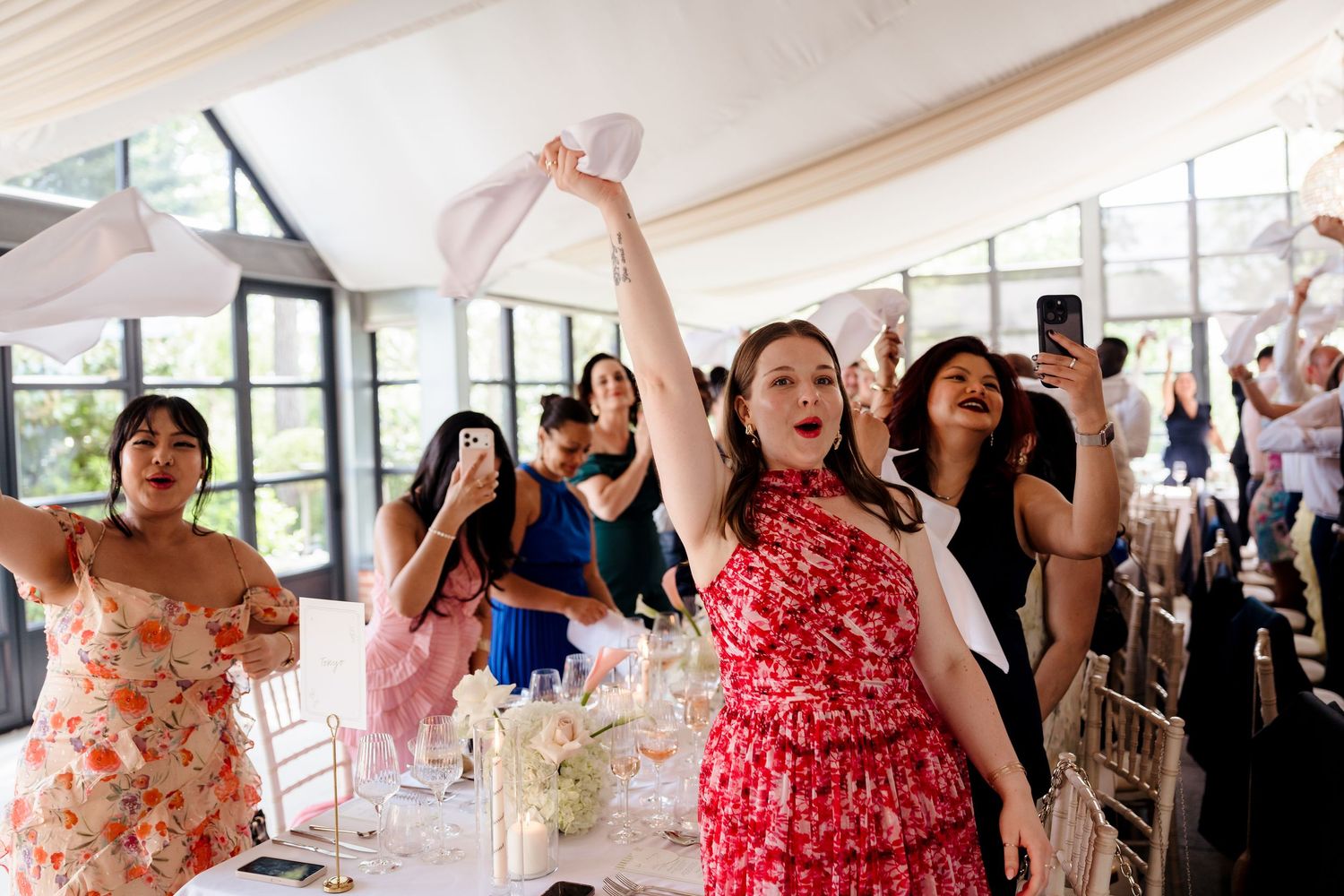Excited wedding guests celebrate inside a marquee reception, waving napkins and cheering joyfully.