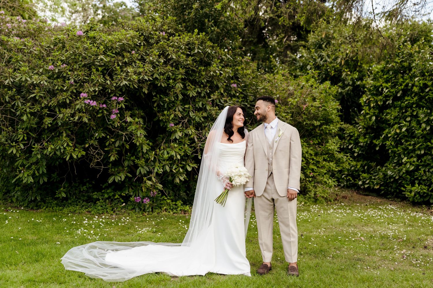 Bride in white gown with veil and groom in beige suit stand together on green lawn with lush garden backdrop.