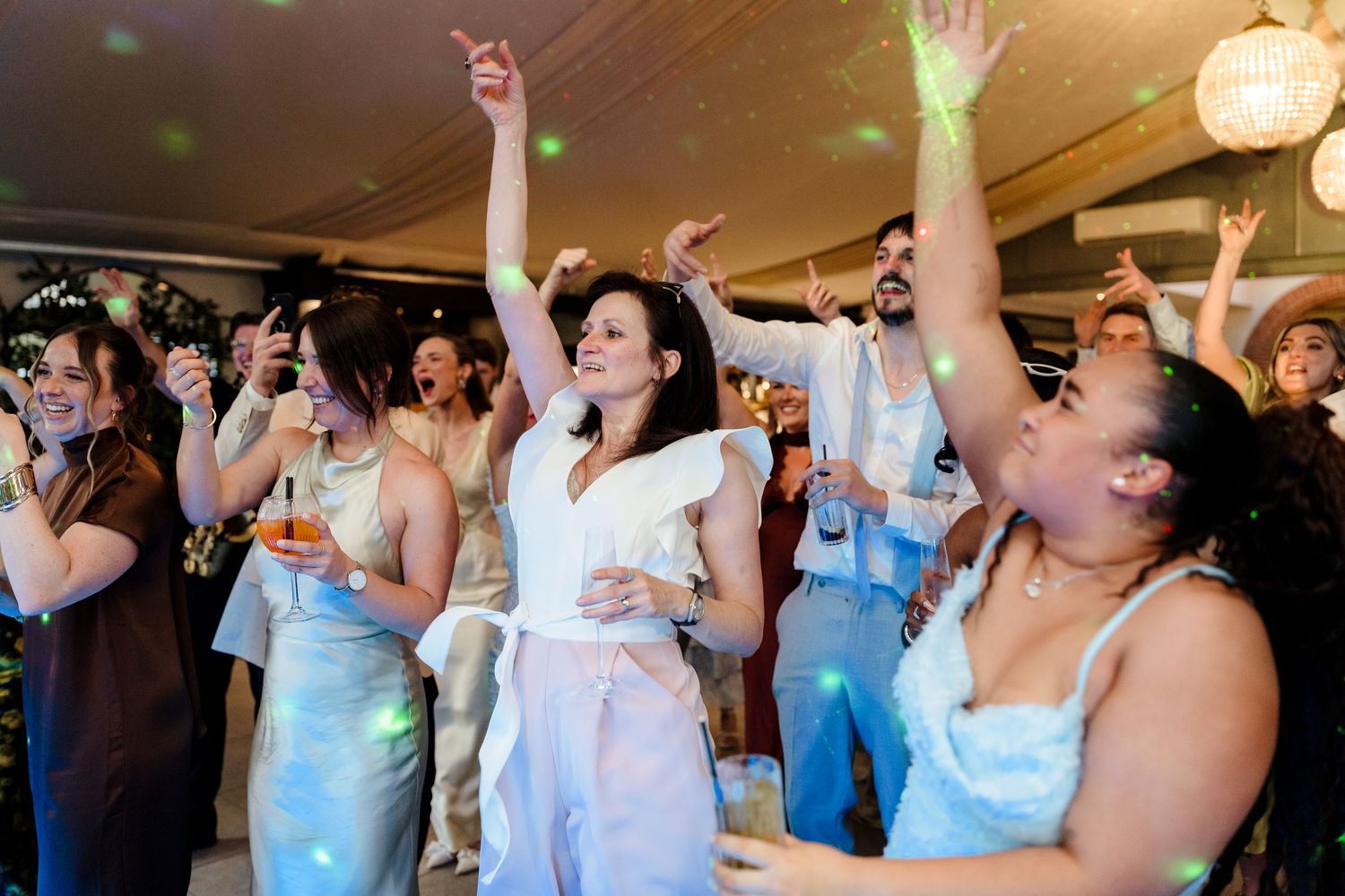 Wedding guests including the bride dance energetically with hands raised under disco lights at reception.