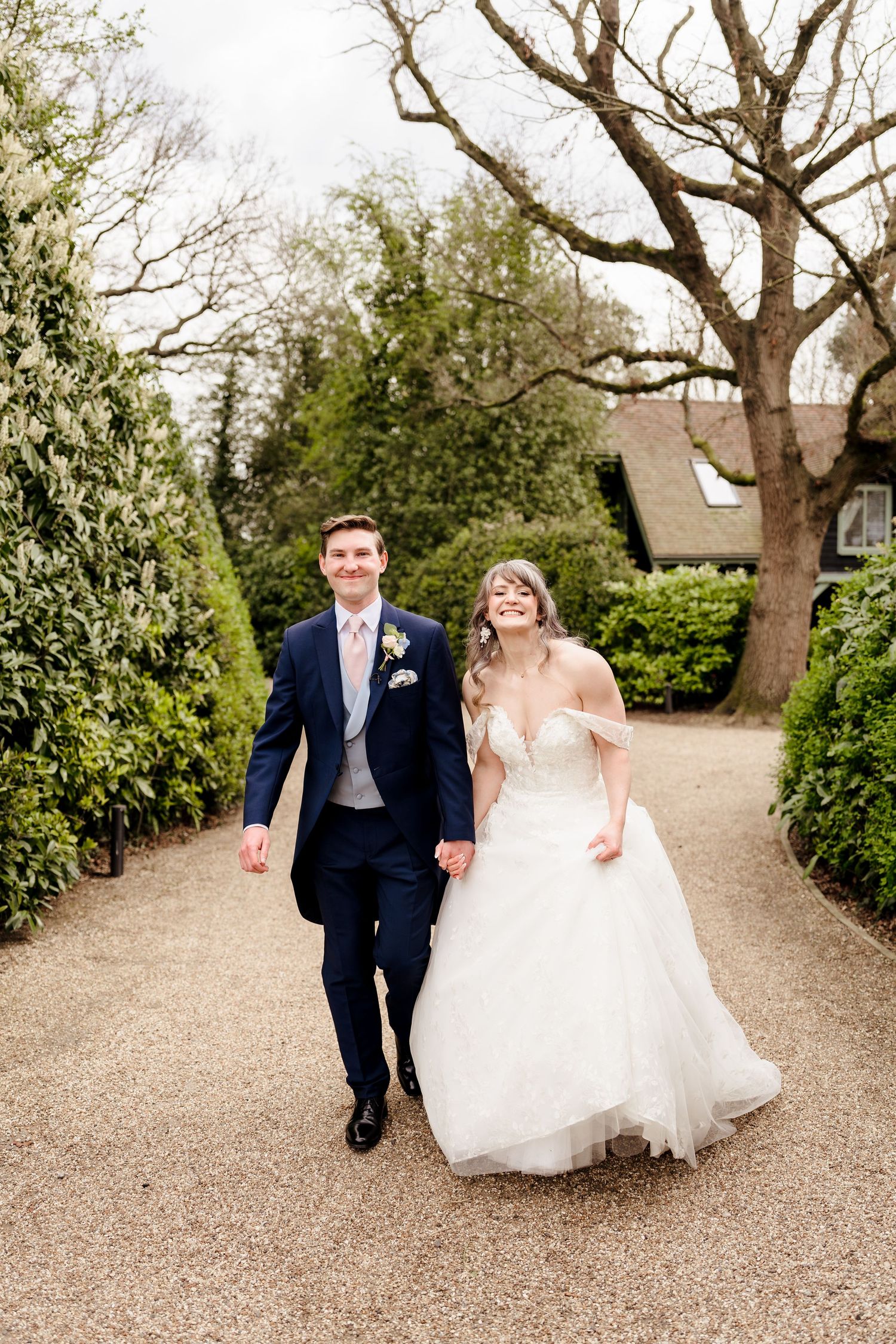 Smiling bride and groom walk hand-in-hand along a garden path on their wedding day.
