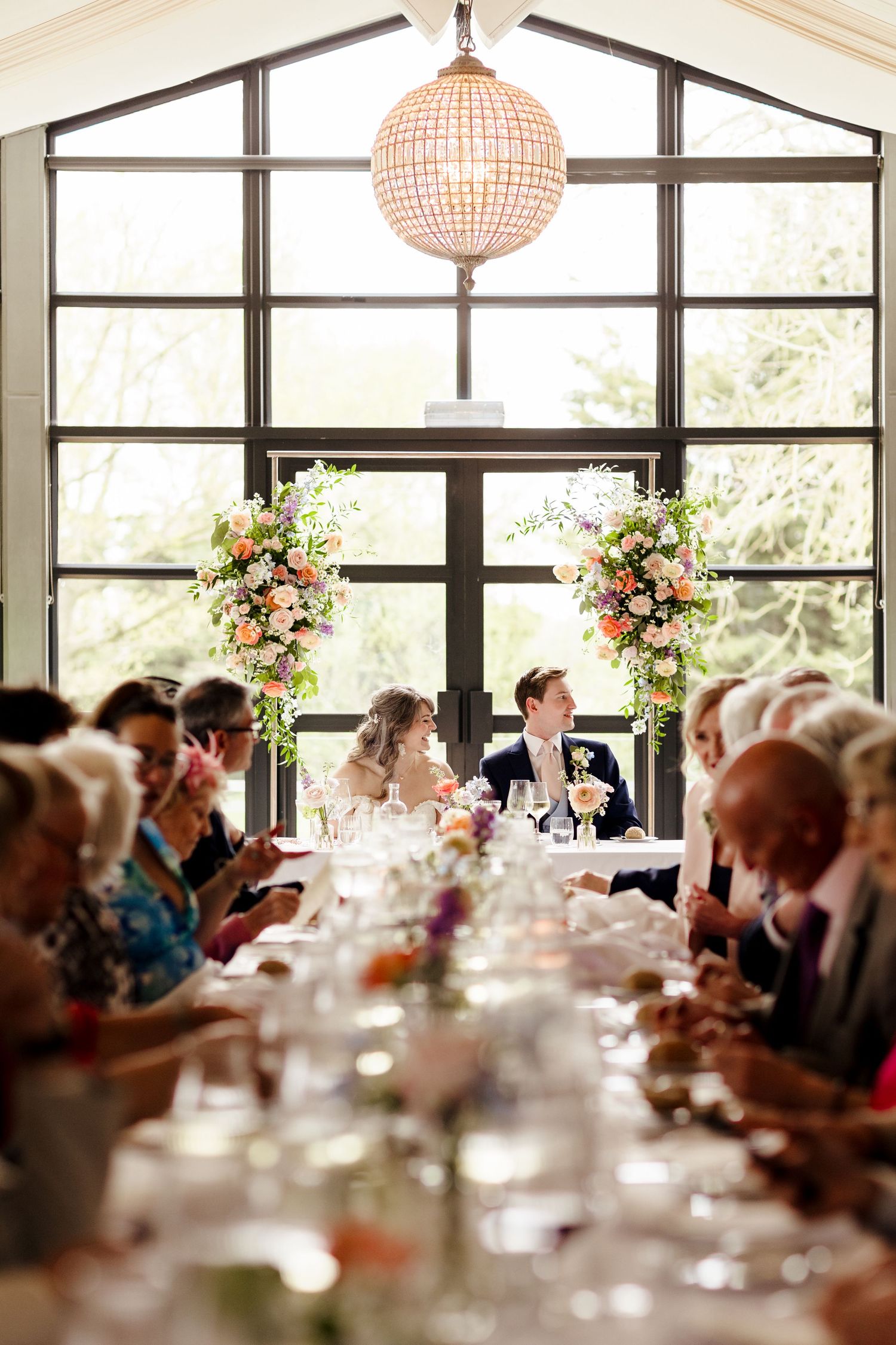 Bride and groom seated at elegant wedding reception table with floral arrangements and chandelier.