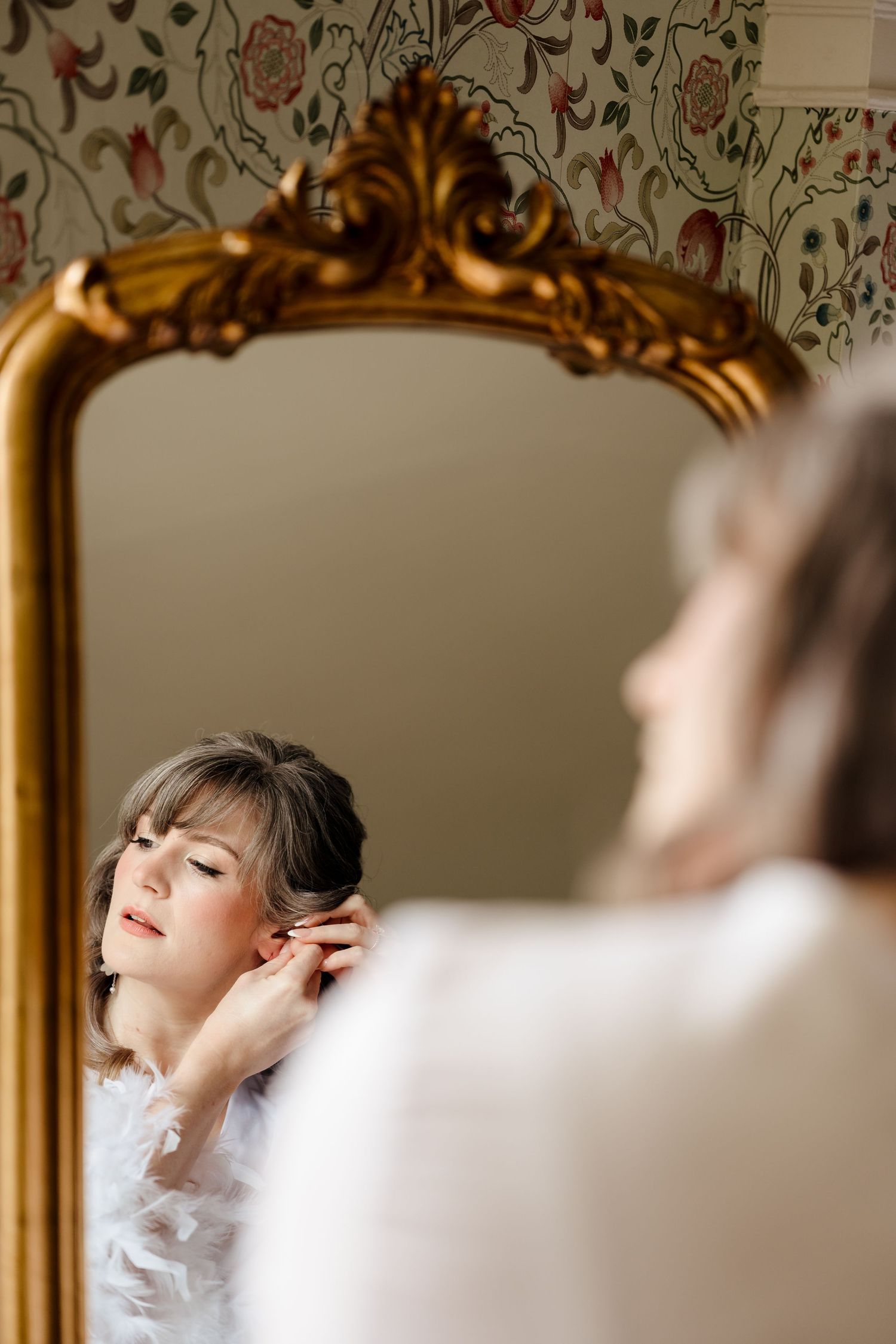 Bride in white robe adjusting earring while gazing into an ornate gold-framed mirror during wedding preparations.