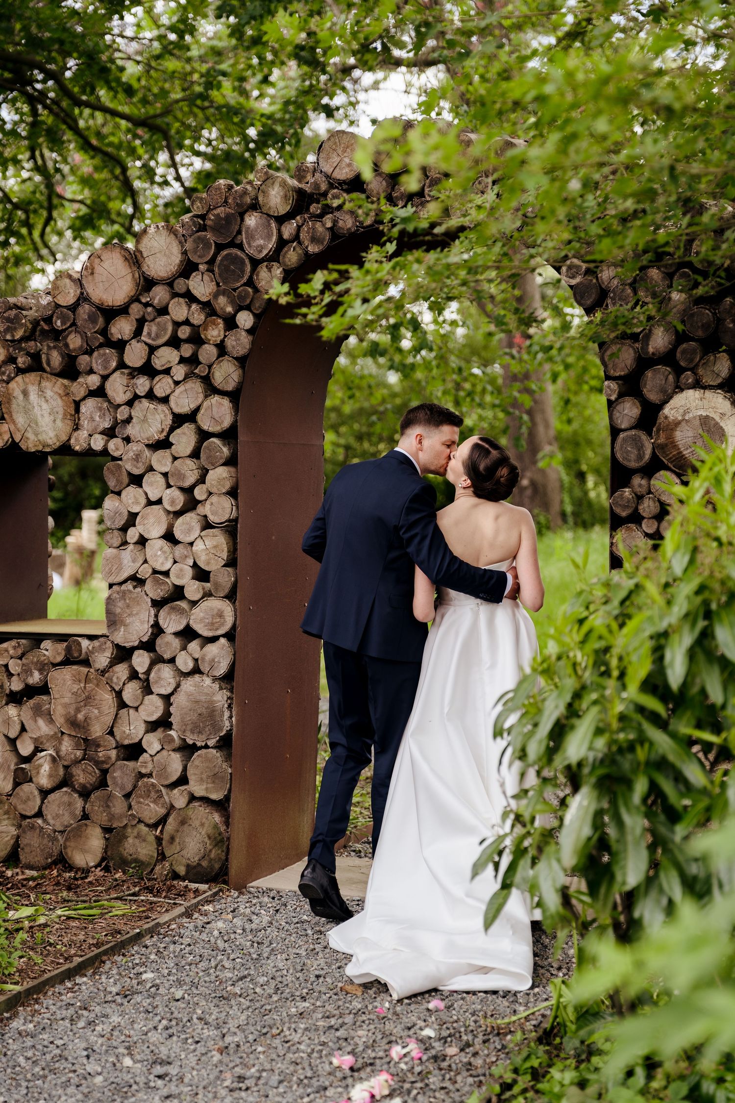 A bride and groom share a romantic kiss under a rustic log arch surrounded by lush green foliage.