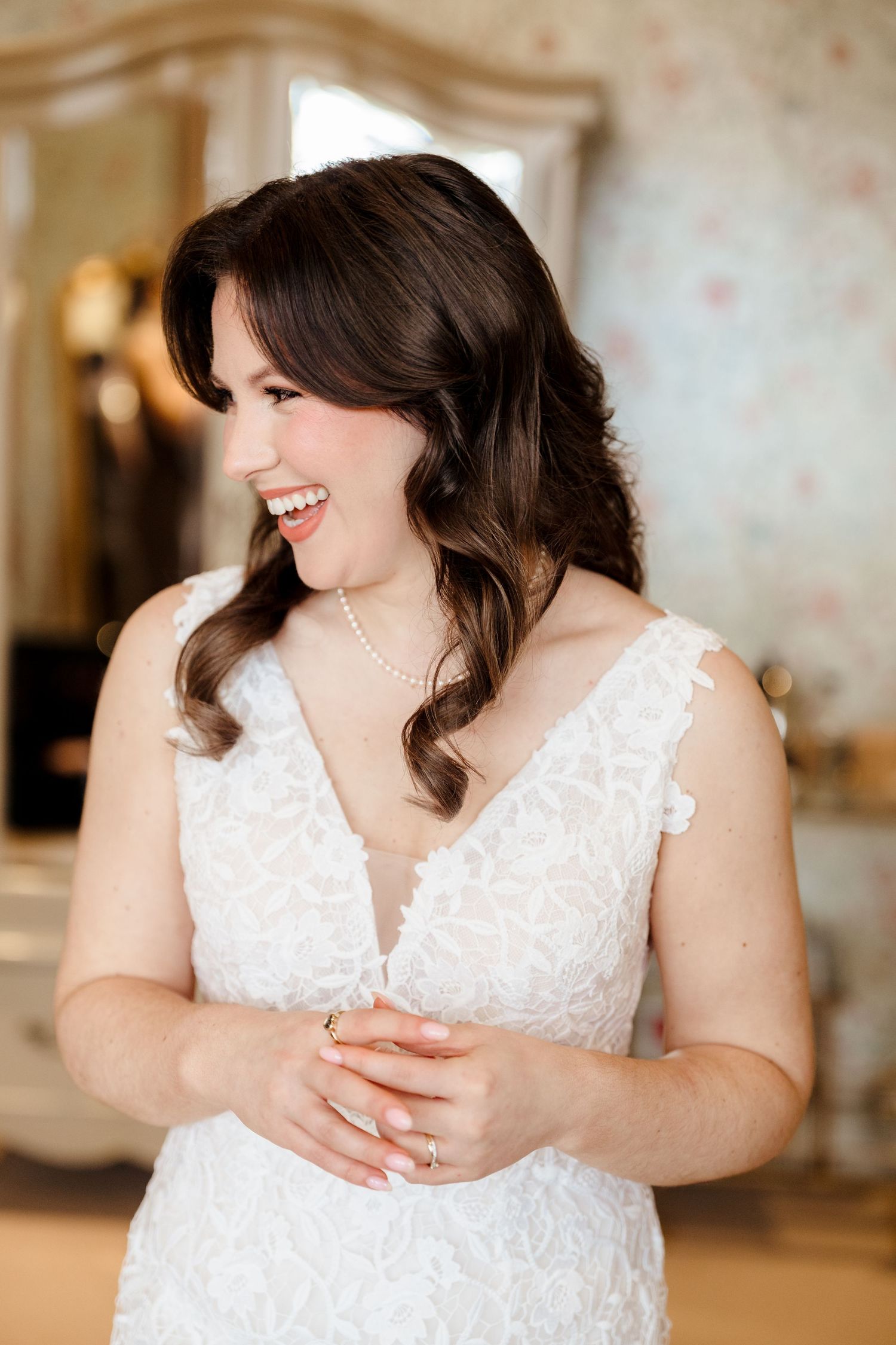 Smiling bride in white lace V-neck wedding dress with pearl necklace and wavy dark hair.