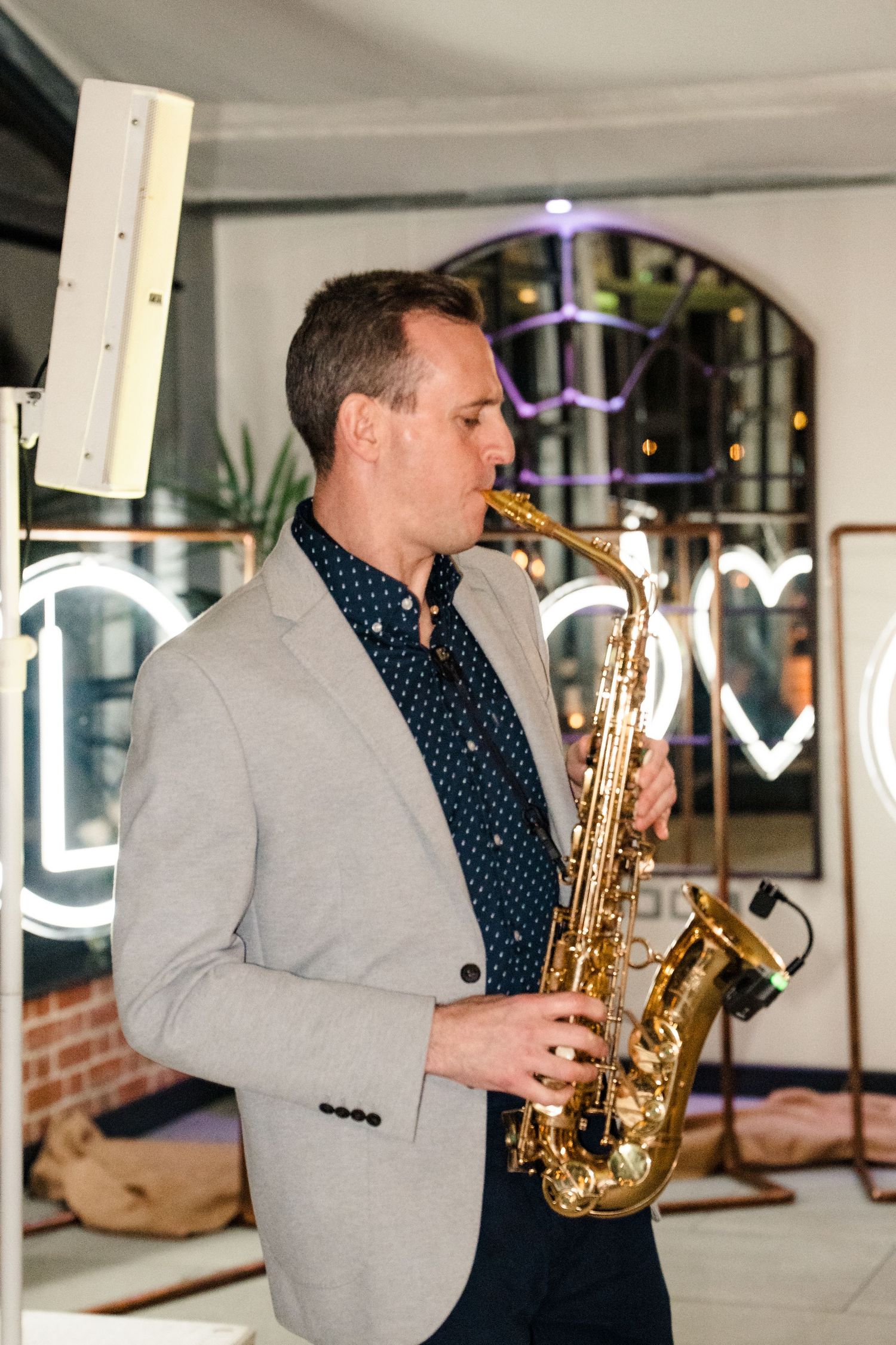 Saxophonist in grey blazer performing at wedding reception with neon LOVE sign in background.