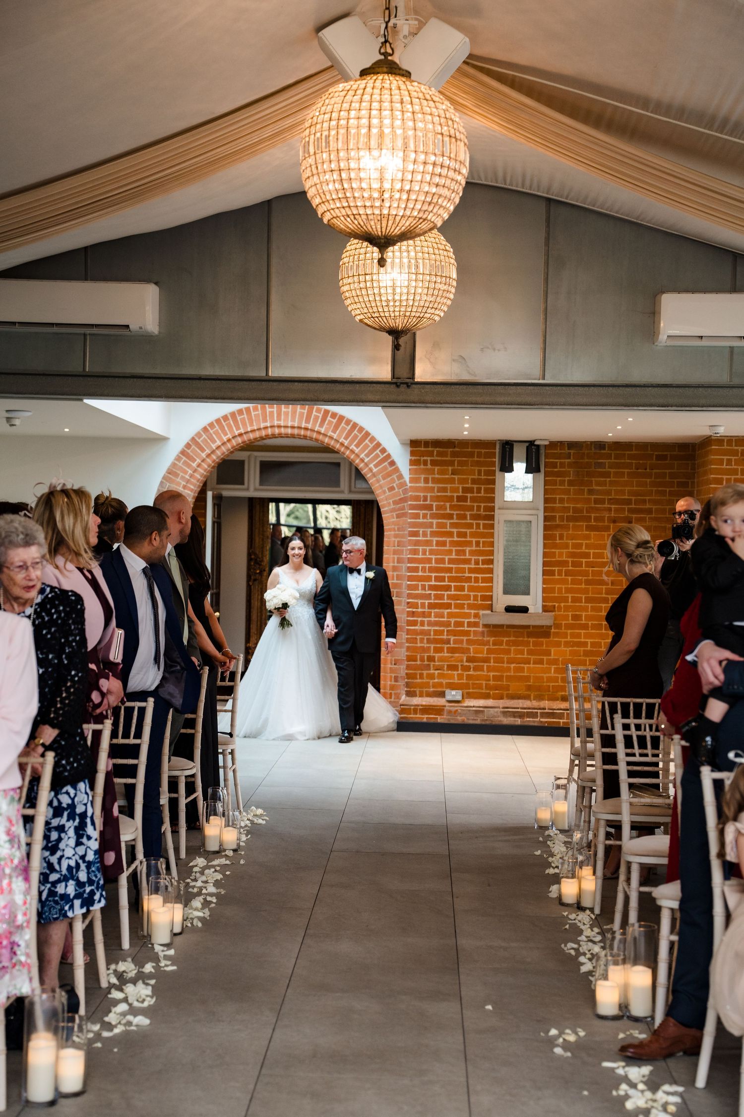 Bride walks down candlelit wedding ceremony aisle with her father in a brick-walled venue.