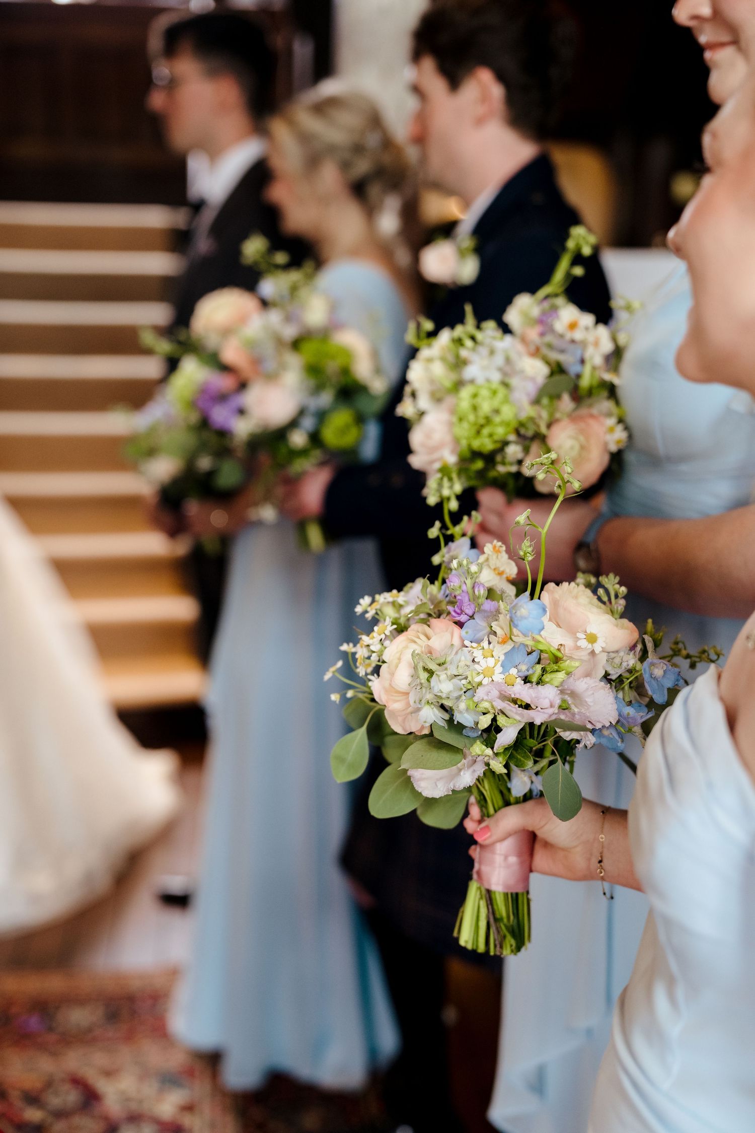 Bridesmaids in light blue dresses holding pastel floral bouquets with roses and greenery at a wedding ceremony.