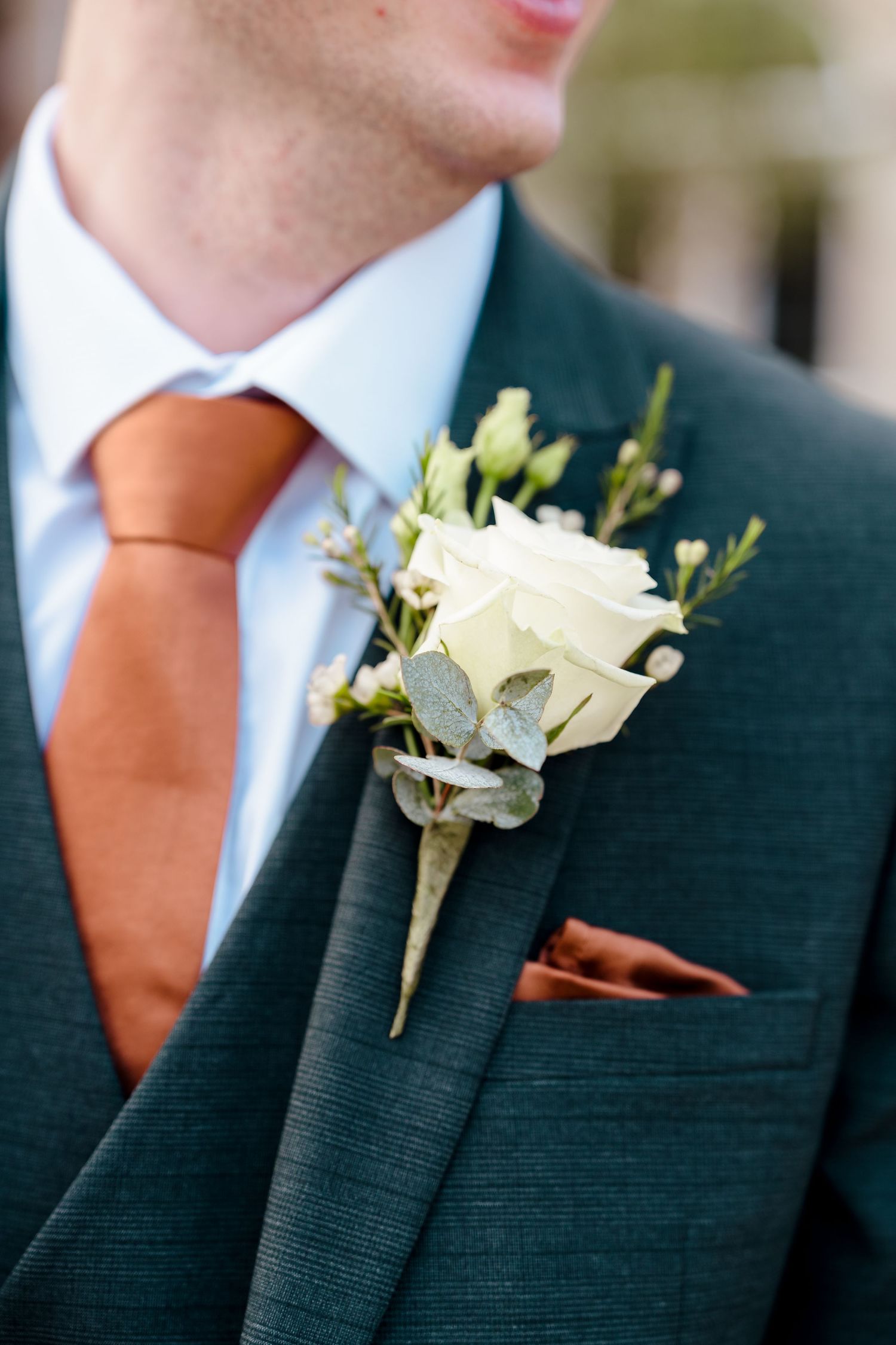 Groom wearing dark suit with white rose boutonniere and burnt orange tie and pocket square at wedding.
