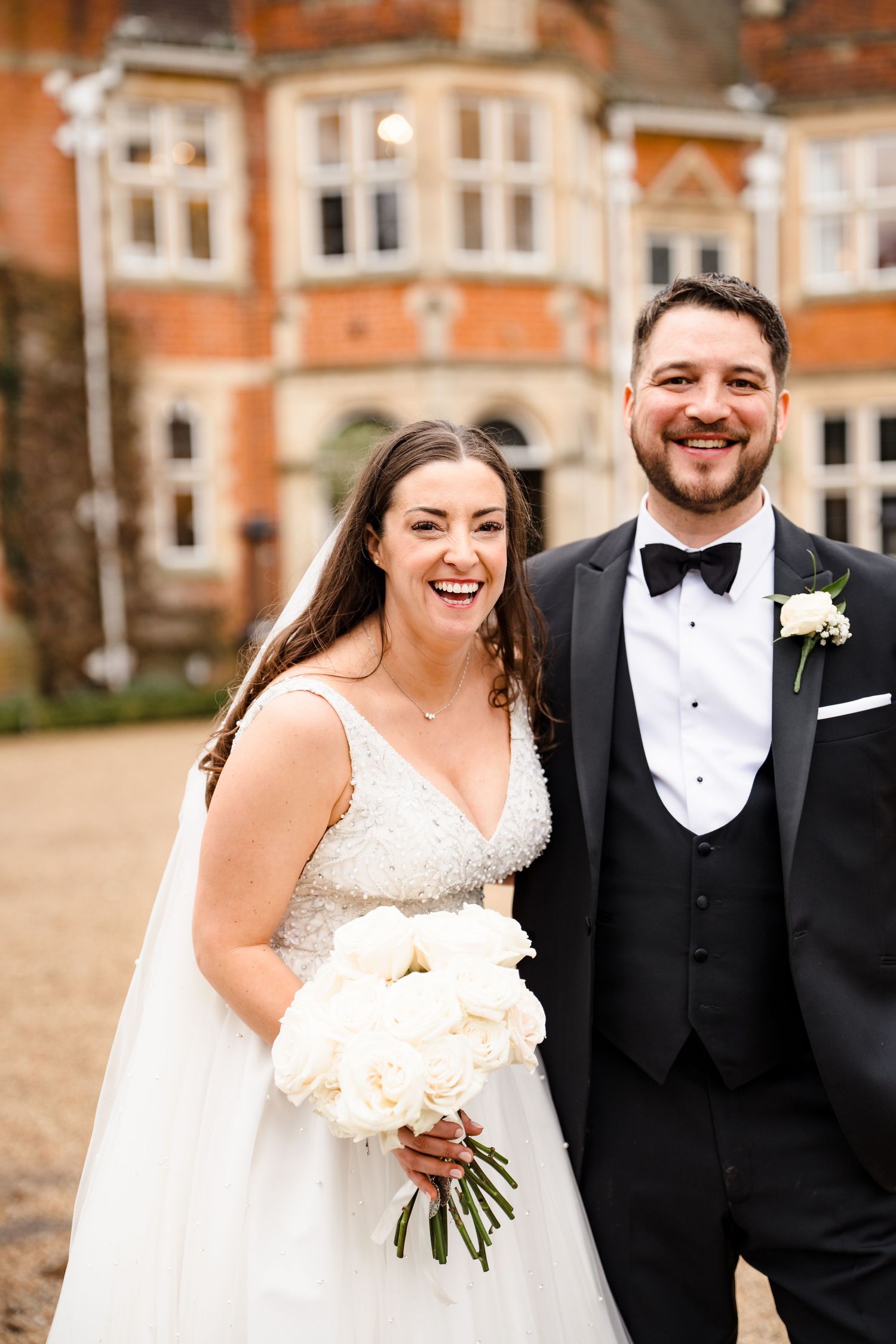 Smiling bride holding white peony bouquet stands with groom in black tuxedo outside elegant brick manor house.