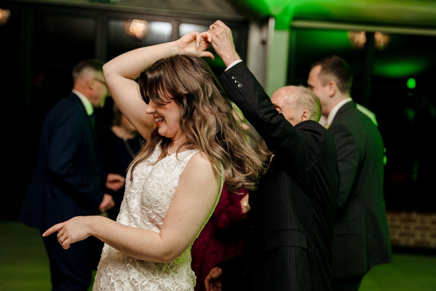 Bride in lace gown spinning on wedding reception dance floor with green ambient lighting.