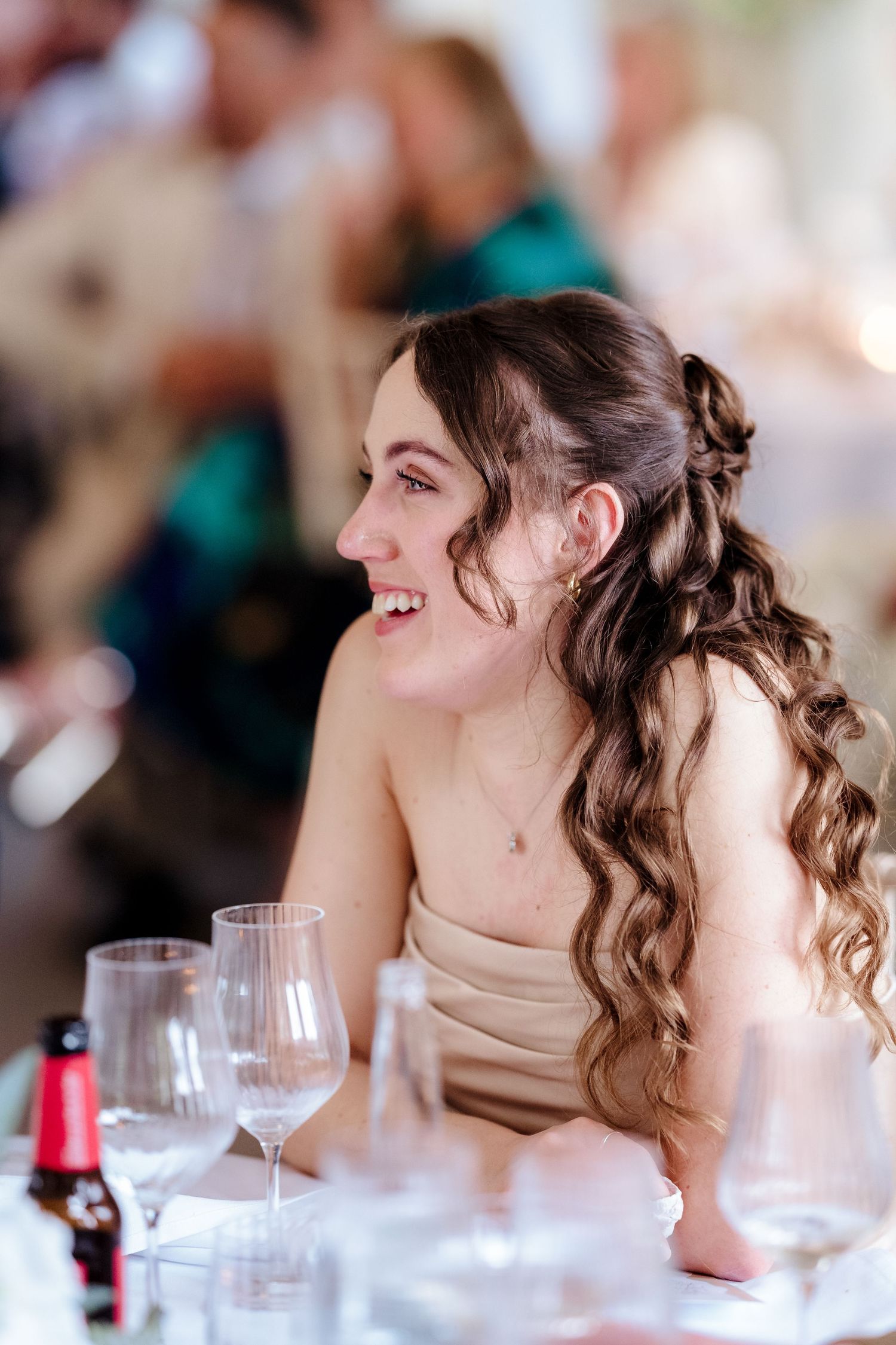 A smiling bridesmaid with curly hair enjoys the wedding reception seated at a decorated table.
