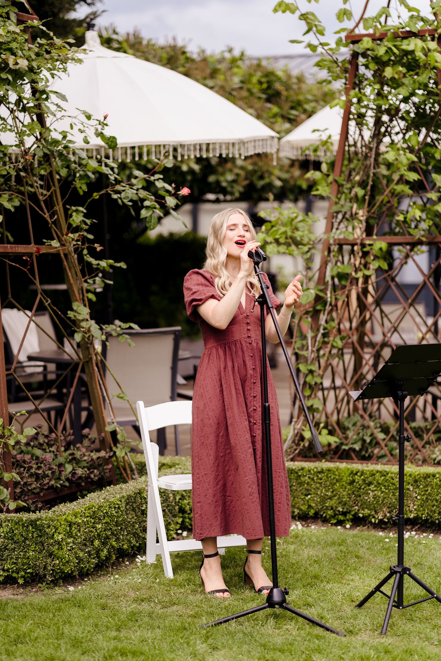 A blonde woman in a burgundy dress sings at a microphone stand in a lush garden wedding venue.