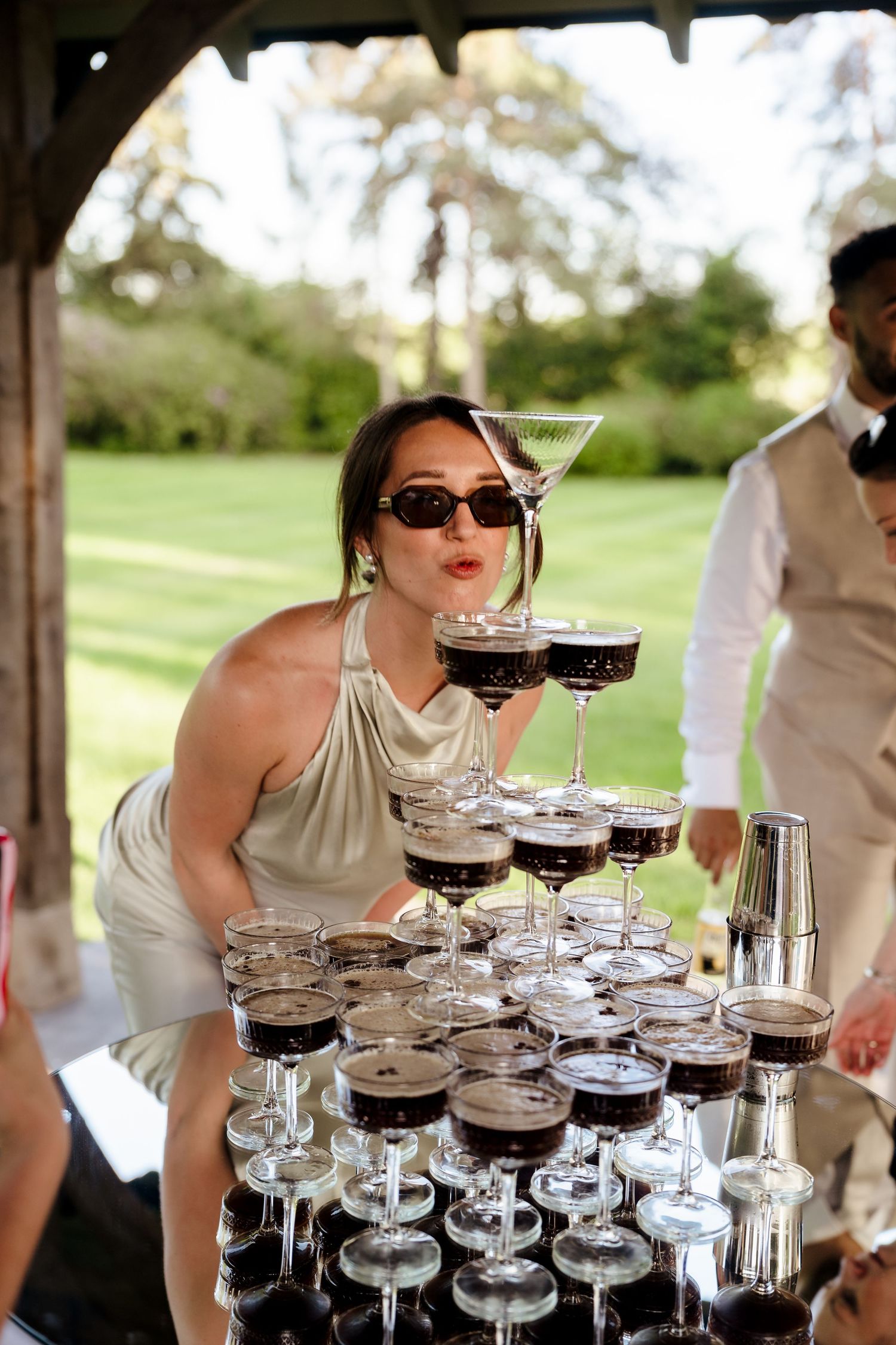 Woman in sunglasses poses playfully behind a tower of espresso martini glasses at an outdoor event.