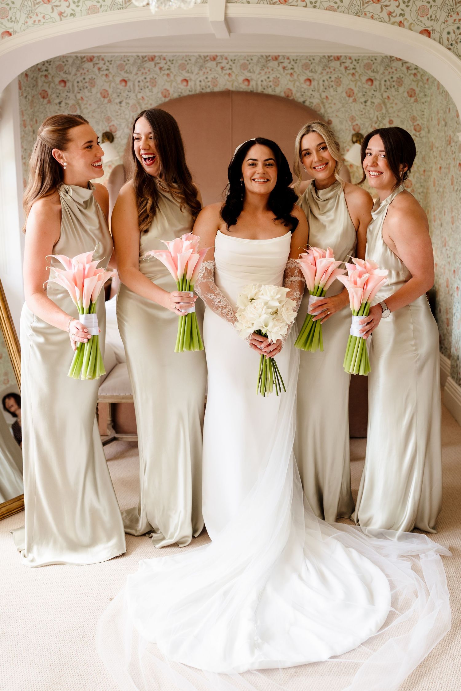 Bride in white gown with four bridesmaids in sage satin dresses holding pink calla lily bouquets indoors.