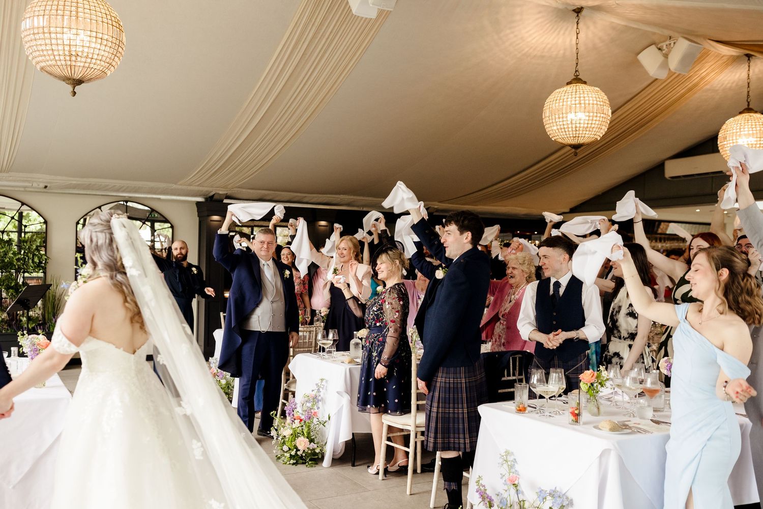 Joyful wedding guests waving napkins as bride and groom enter elegant tented reception venue with chandelier lighting.