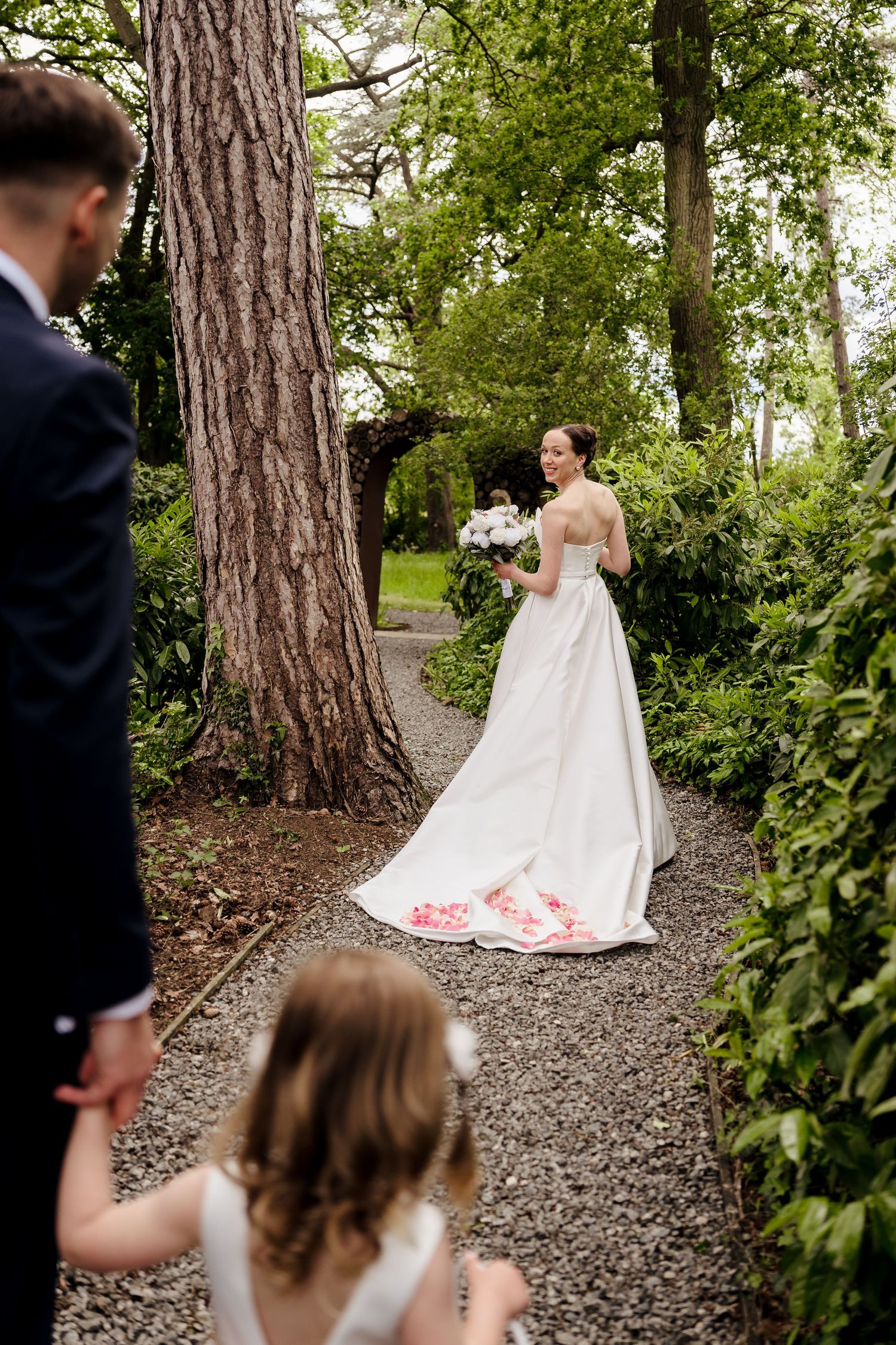 Bride in white gown with floral details holds bouquet on garden path with groom and flower girl.