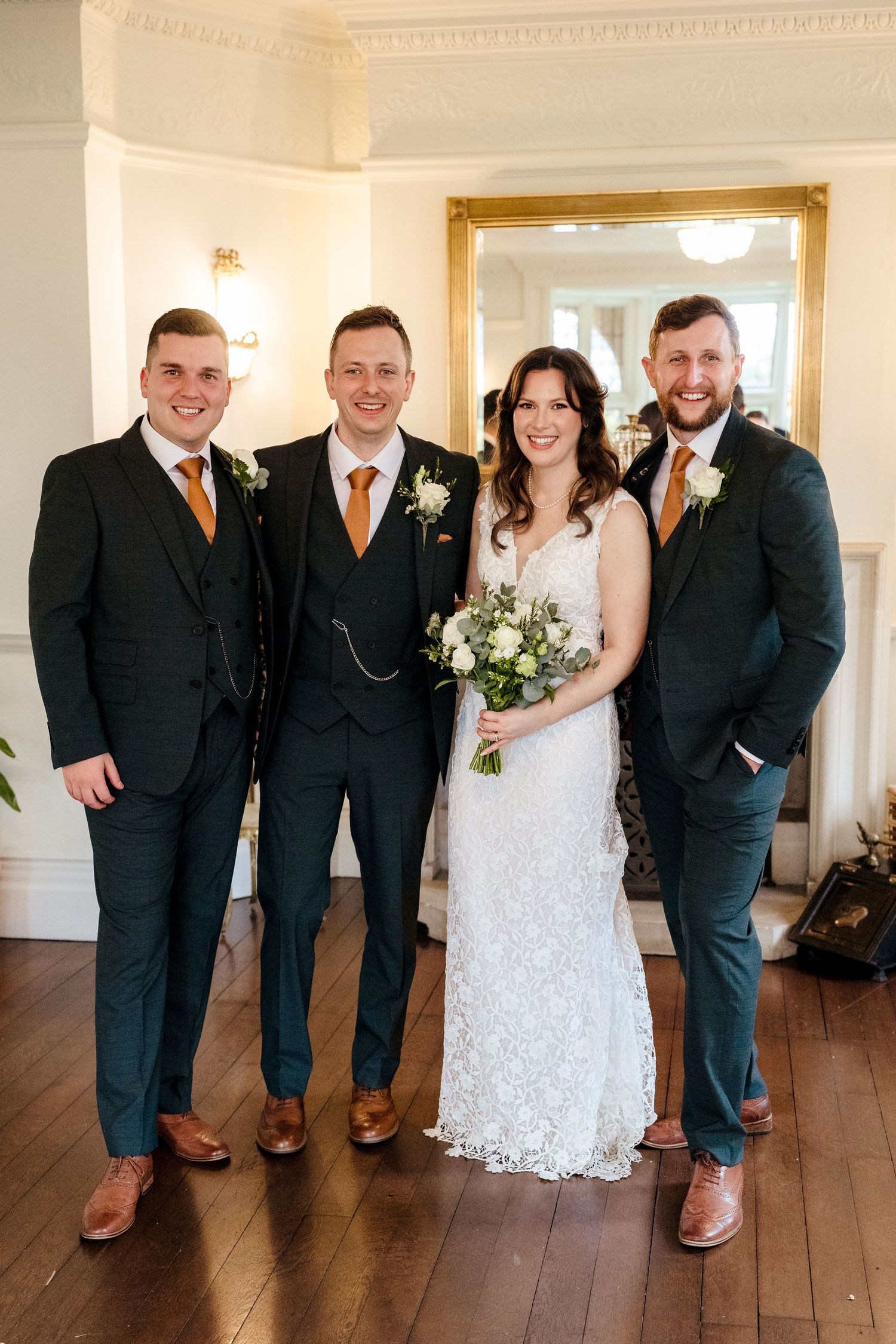 Bride in lace gown poses with groom and two groomsmen in dark suits with orange boutonnieres indoors.