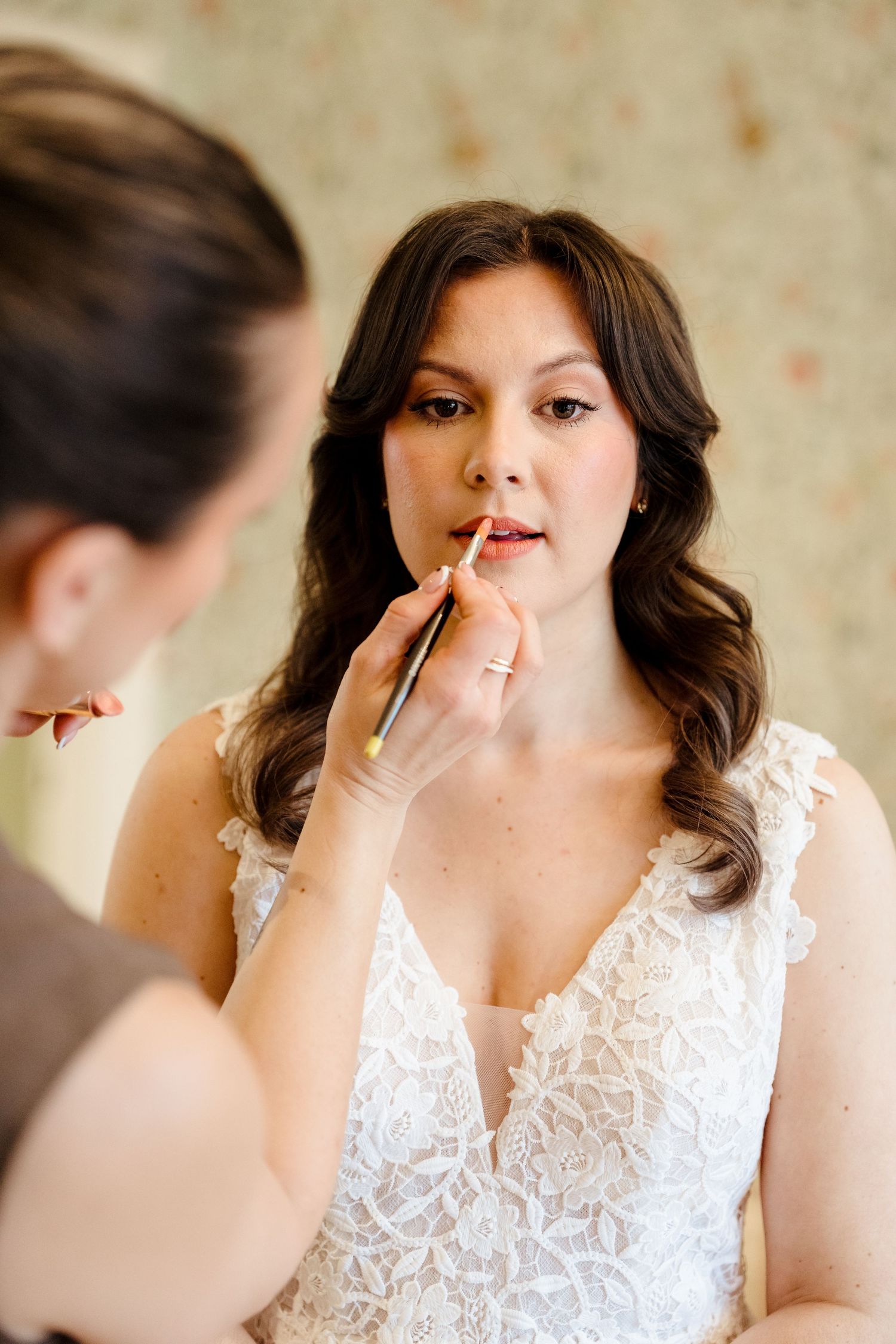 Makeup artist applying lipstick to a bride in a white lace dress on her wedding day.