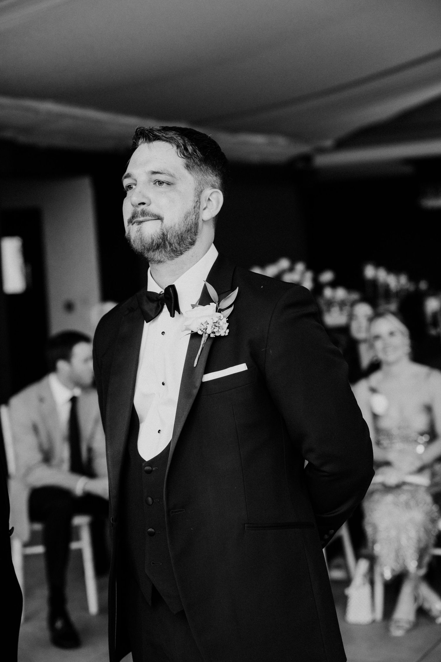 Groom in black tuxedo with bow tie and boutonniere waits at wedding altar in black and white photo.