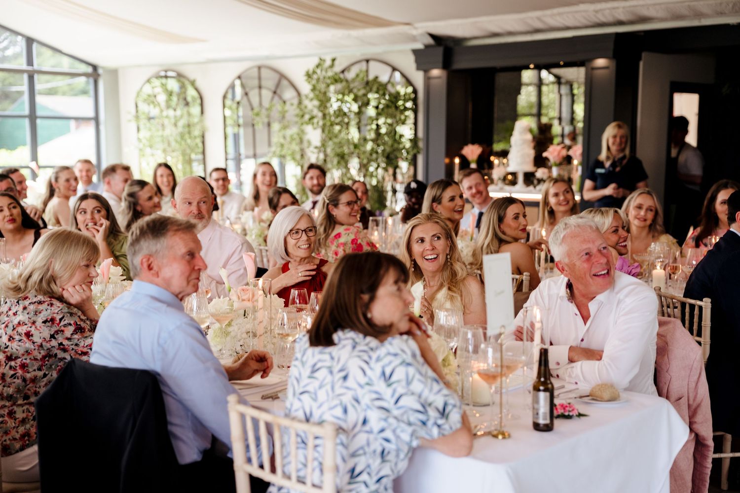 Wedding guests laugh and chat at round tables in bright glass-roofed reception venue with champagne.