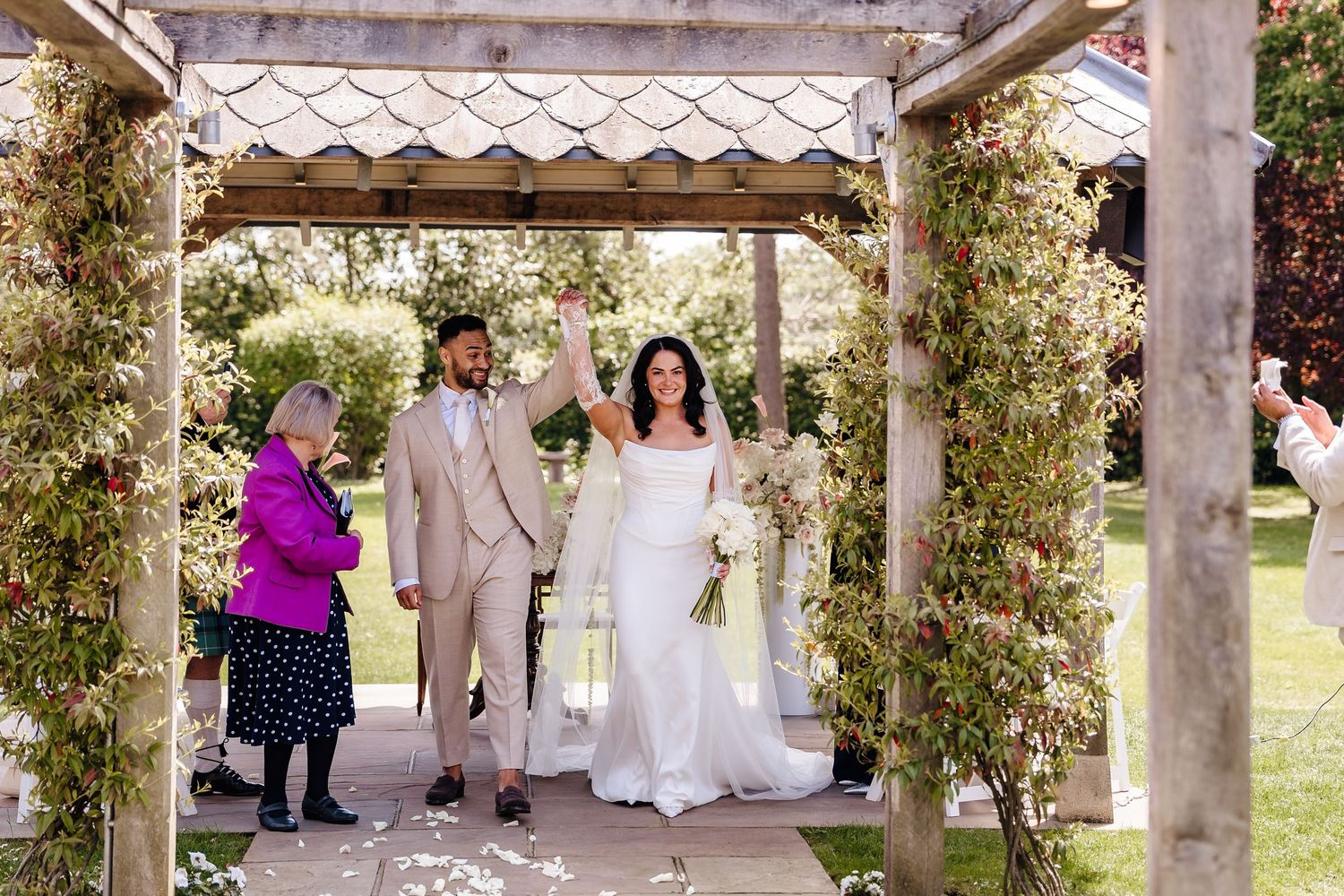 Bride and groom celebrate under a floral pergola at an outdoor wedding ceremony with a guest in purple nearby.