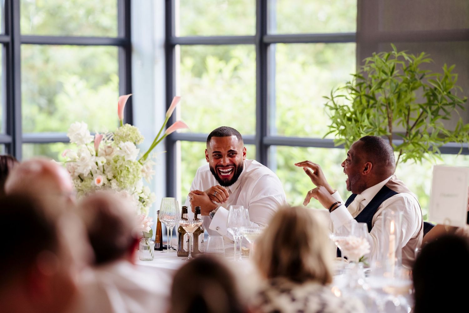 Two wedding guests laugh joyfully together at a decorated reception table with floral centerpieces.