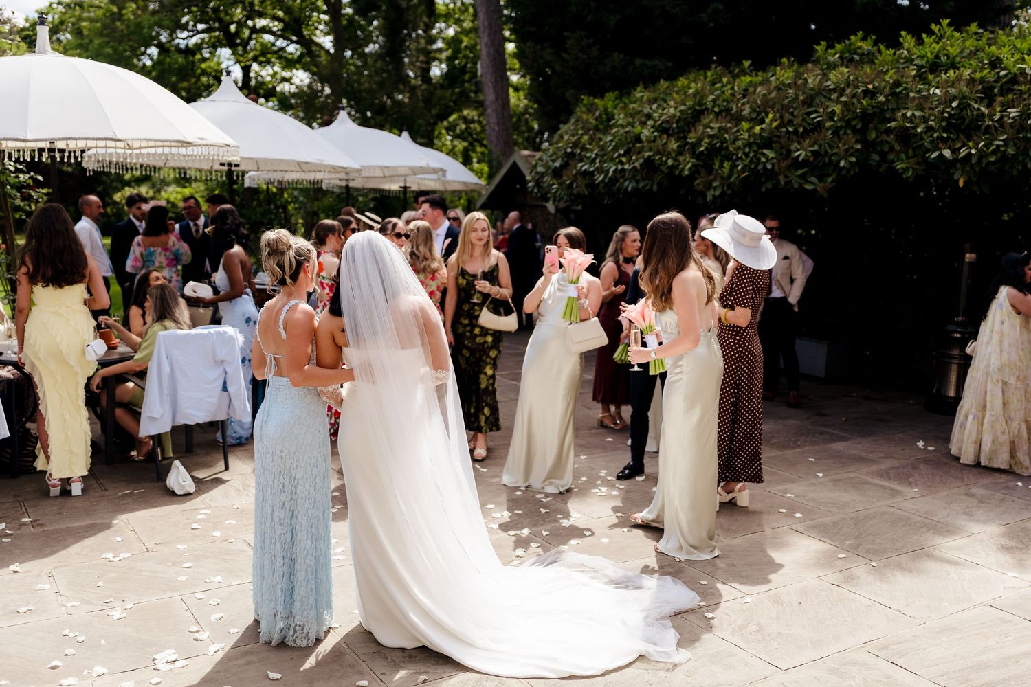 Bride in white gown and veil surrounded by bridesmaids in neutral dresses at outdoor wedding.
