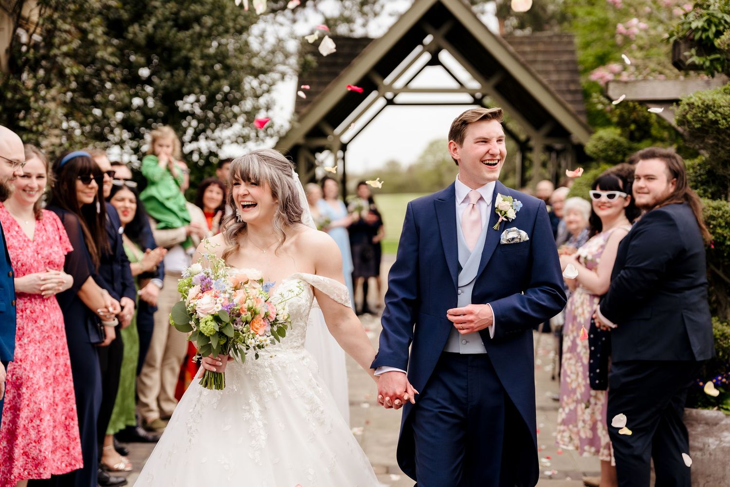 Bride and groom walking through confetti shower with guests celebrating at outdoor wedding ceremony.