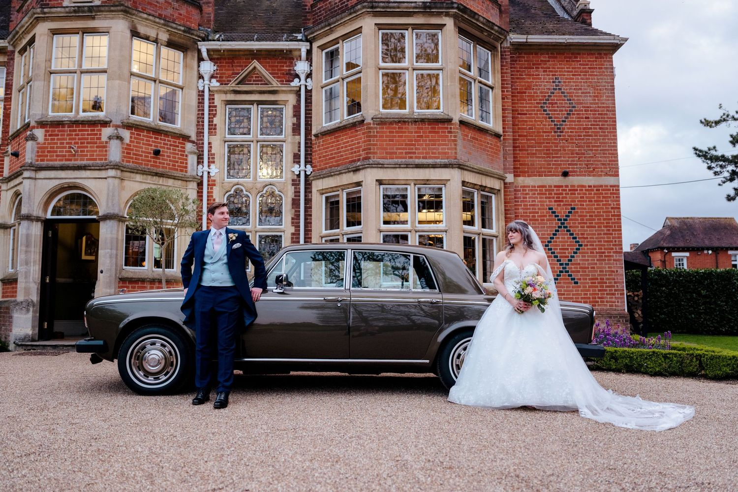 Bride and groom pose beside vintage Rolls-Royce in front of elegant red brick manor.