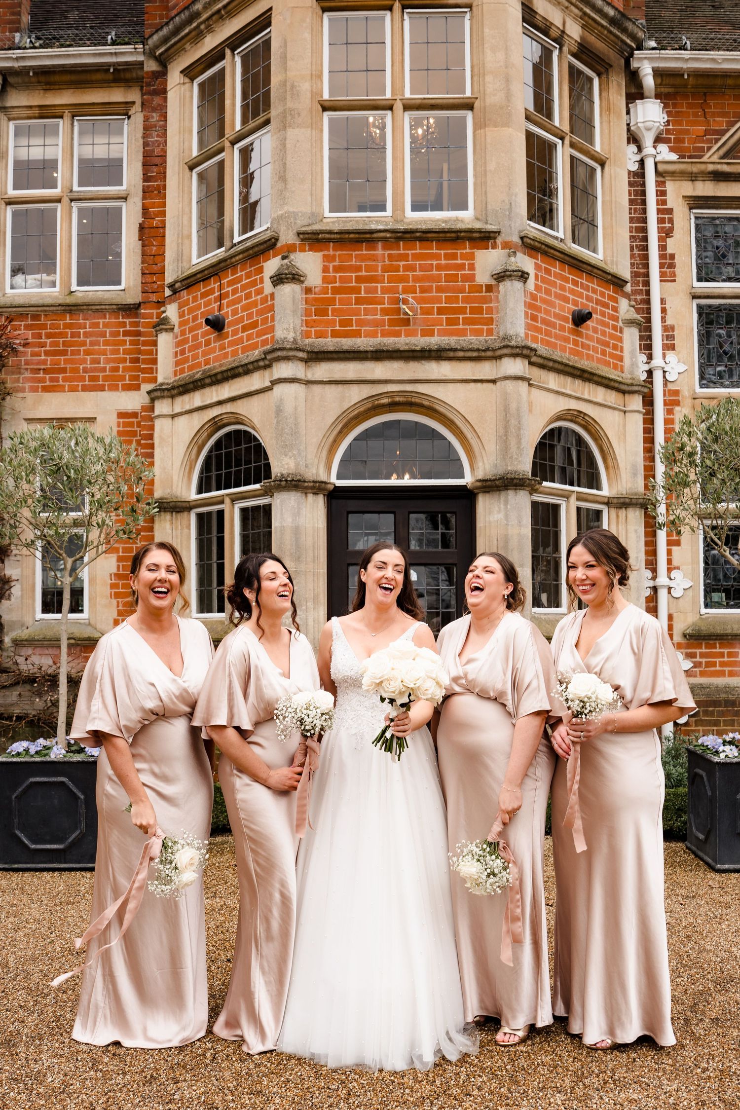 Bride with four bridesmaids in champagne satin dresses laughing outside a red brick manor house.