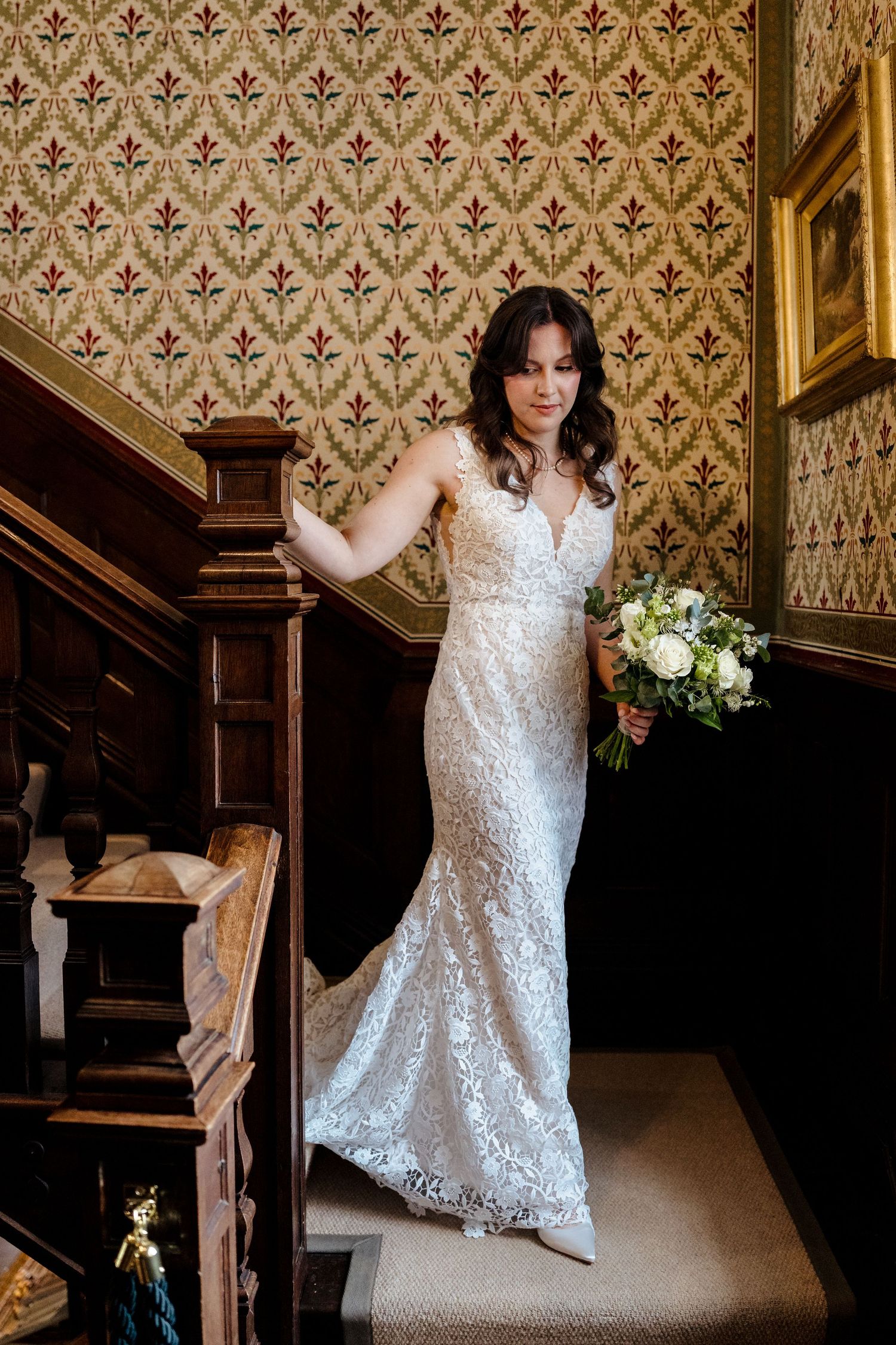 Bride in fitted lace wedding gown holding white bouquet on ornate staircase.