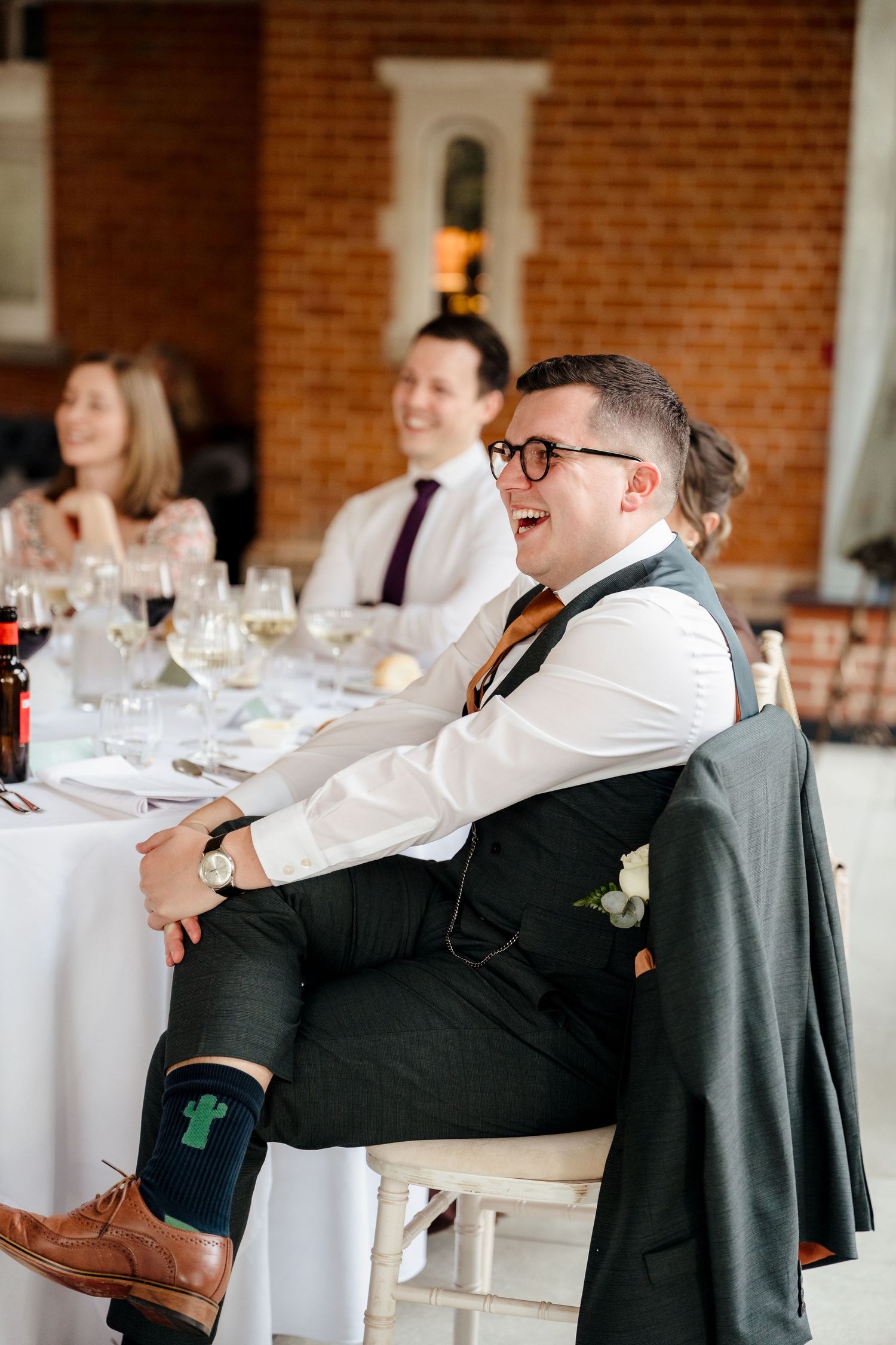 Laughing groomsman in dark suit with cactus socks sits relaxed during wedding reception speeches at brick venue.