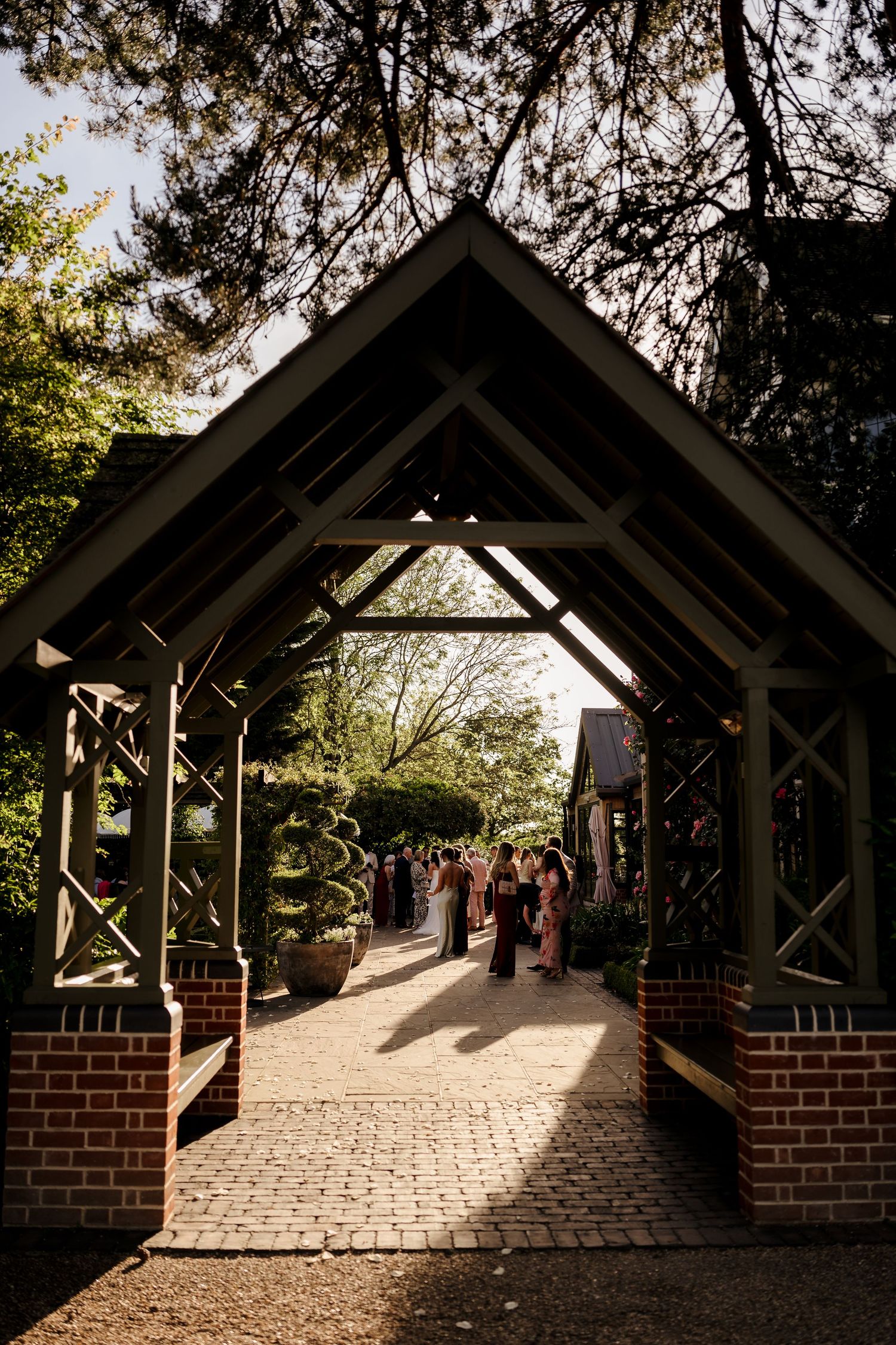 Guests gather in golden hour sunlight beneath a rustic wooden pergola entrance at a wedding venue.