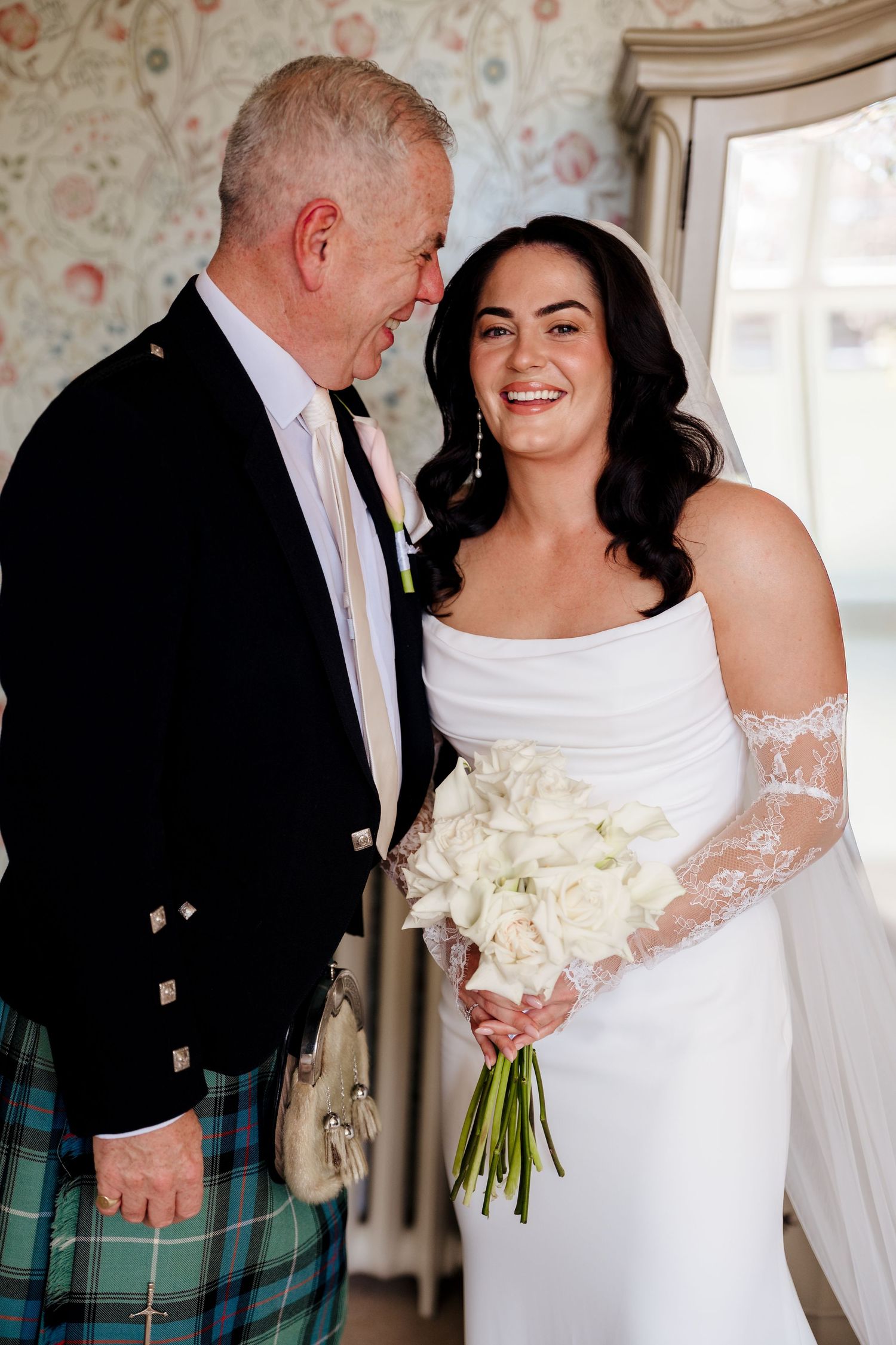Bride in white off-shoulder gown with lace gloves holds white calla lily bouquet beside man in Scottish tartan kilt.