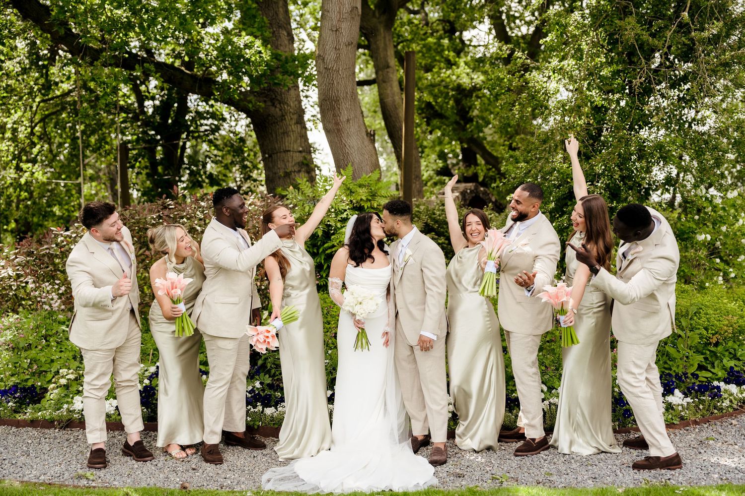 Wedding party celebrating in lush garden, bride and groom kissing surrounded by cheering attendants.