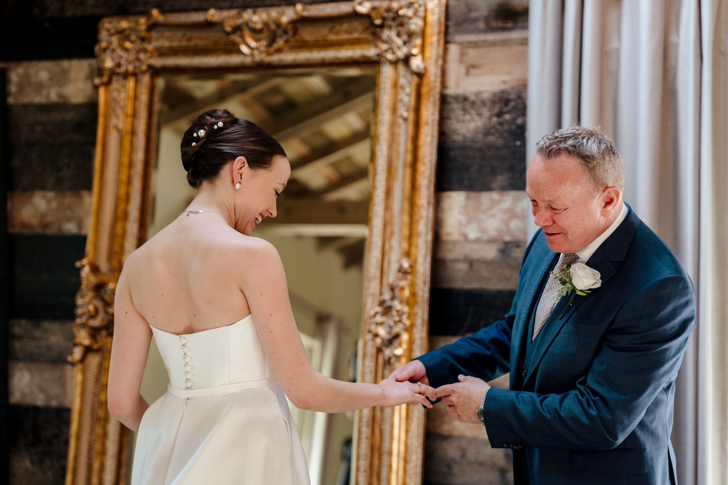 Groom placing wedding ring on bride's finger in front of ornate gold mirror in rustic venue.