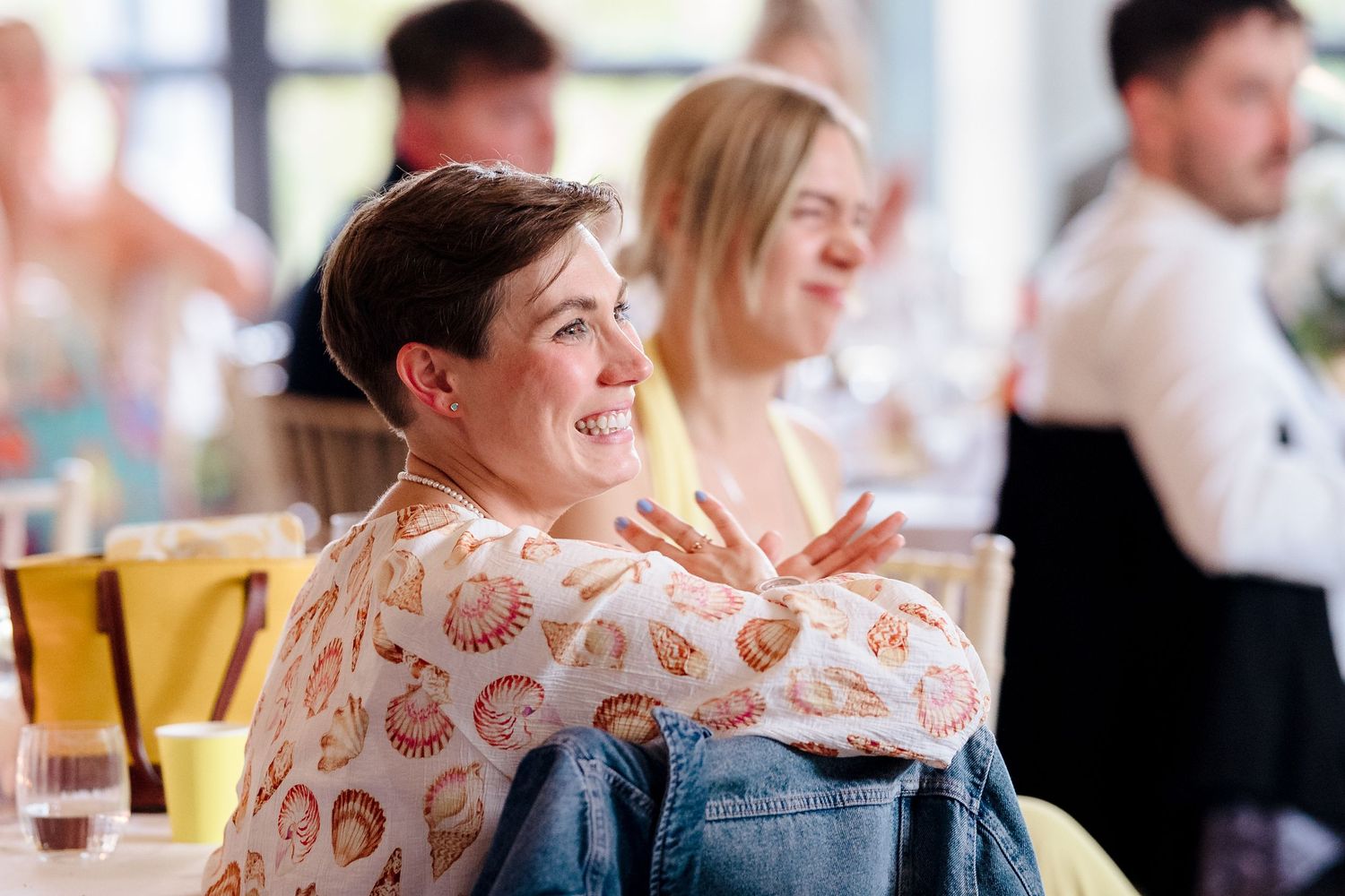 Wedding guest with short hair smiles and claps with arms crossed at joyful reception celebration.