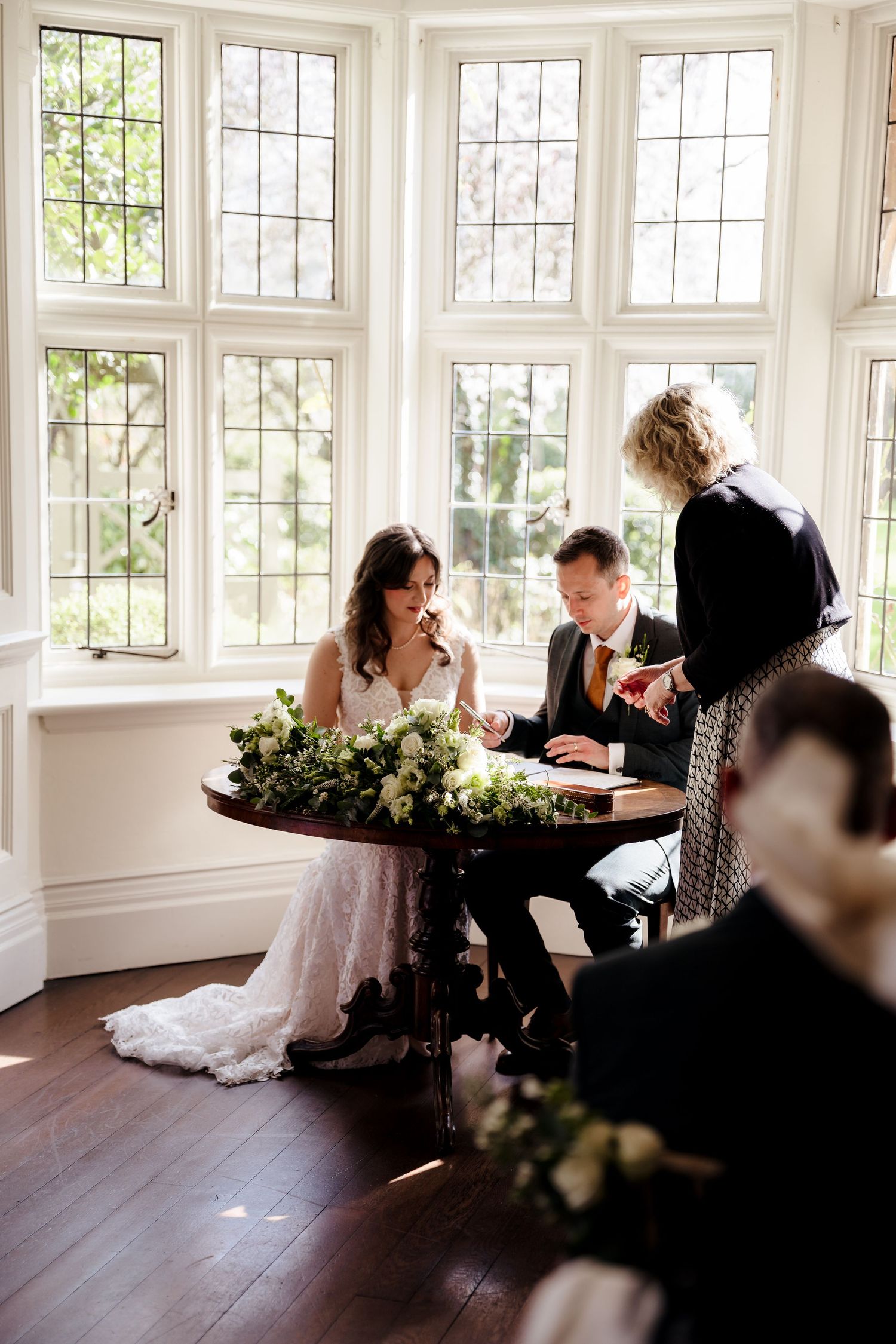 Bride and groom signing wedding register at ornate table adorned with white floral arrangements by large windows.