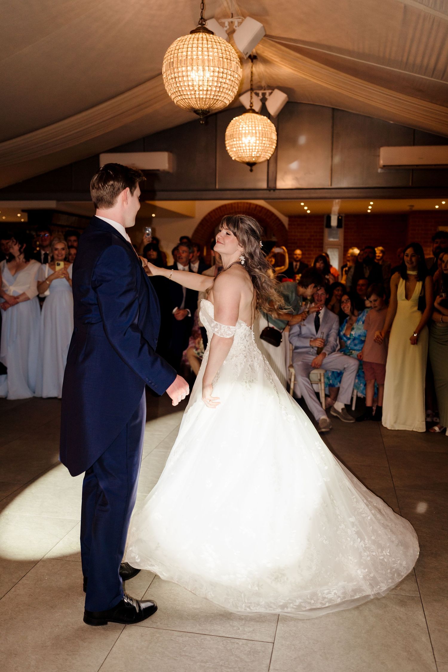 Bride and groom sharing first dance under crystal chandeliers surrounded by wedding guests.