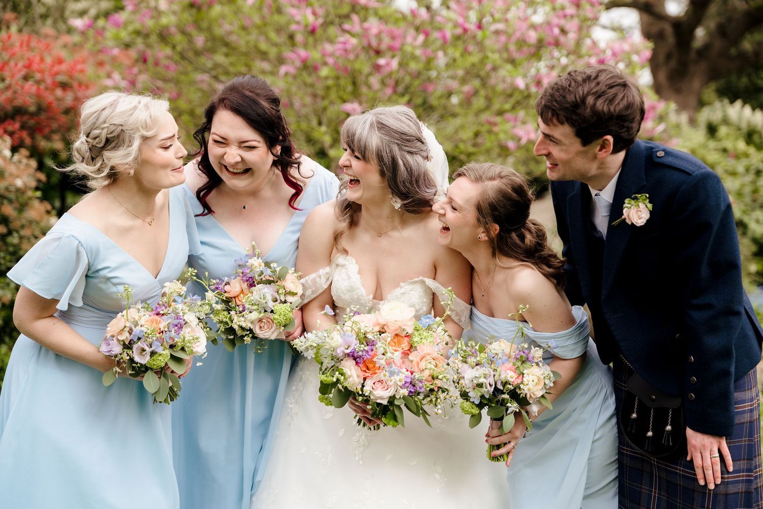 Bride laughing with three bridesmaids in light blue dresses holding colorful bouquets outdoors.