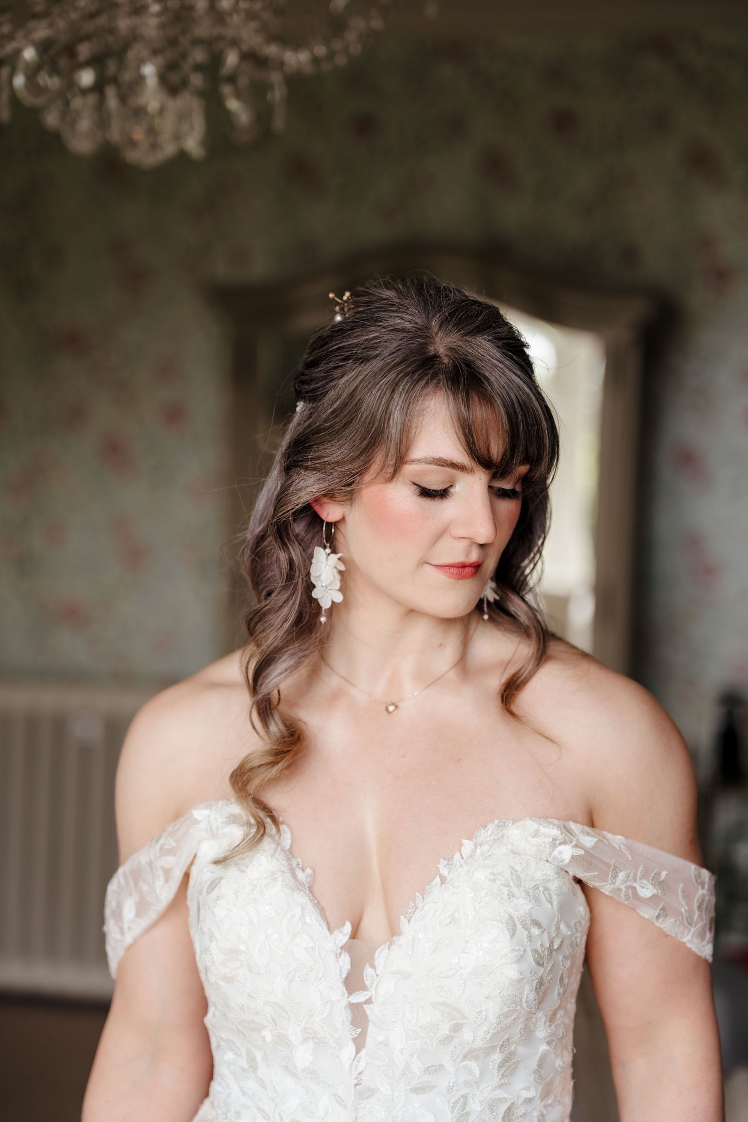 Elegant bride in off-shoulder lace gown with floral earrings beneath crystal chandelier.