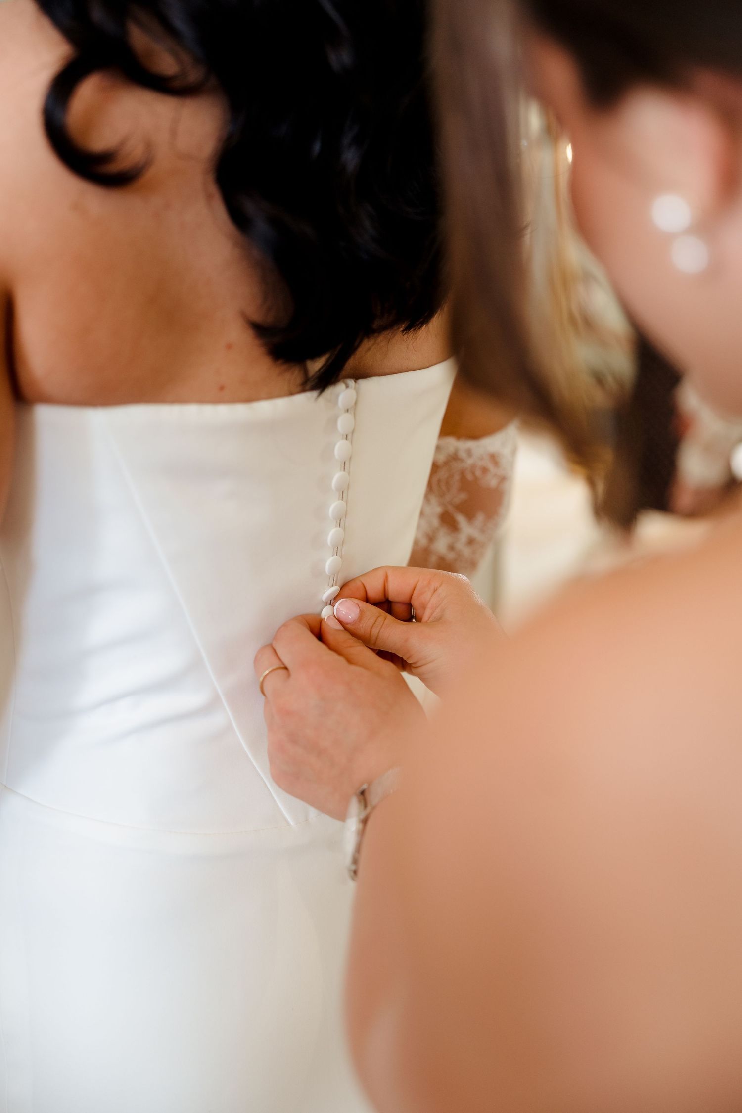 Bridesmaid carefully buttoning the back of a white wedding dress, helping the bride prepare for her special day.