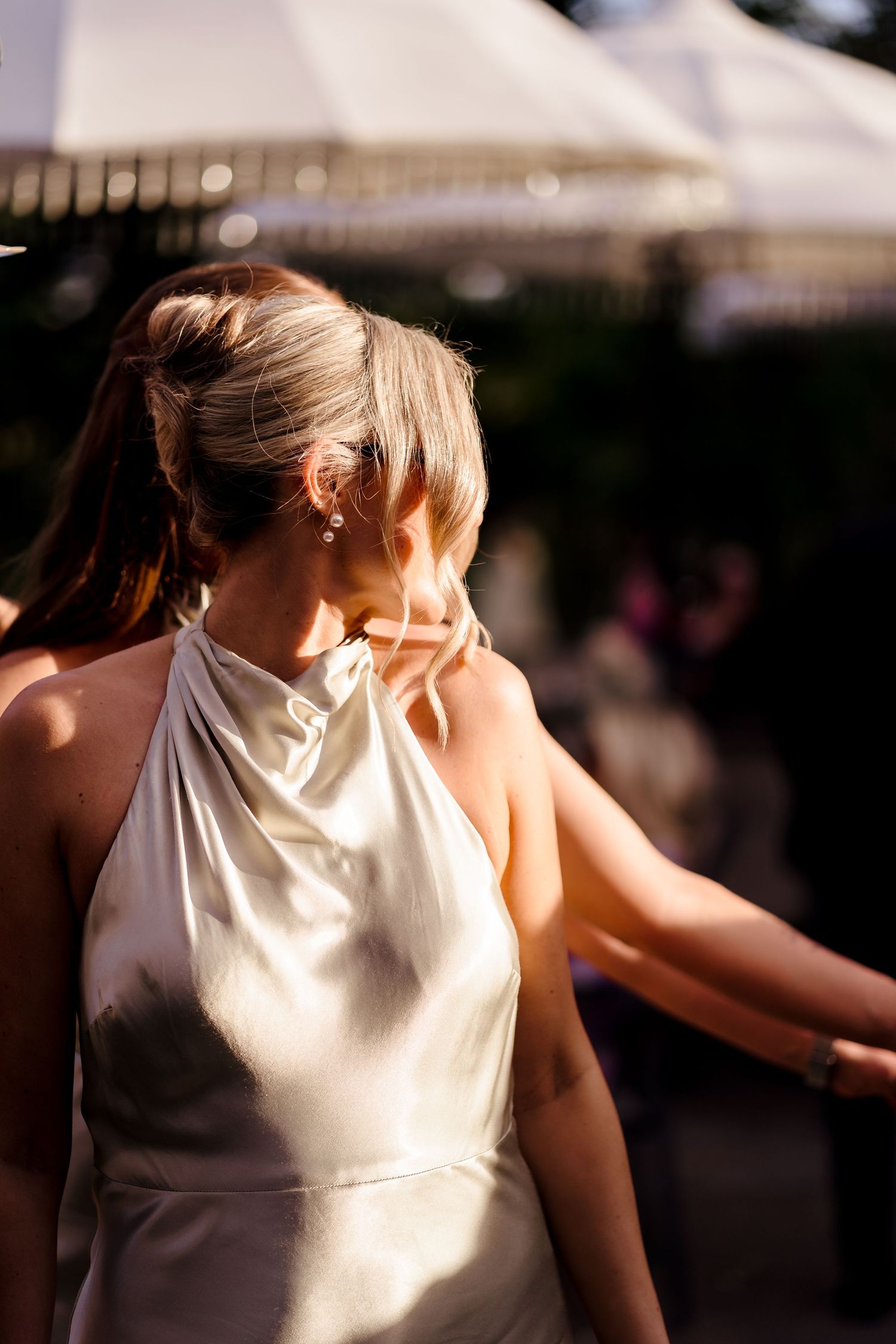 Bridesmaid in ivory halter satin gown with elegant updo earrings in warm golden sunlight outdoors.